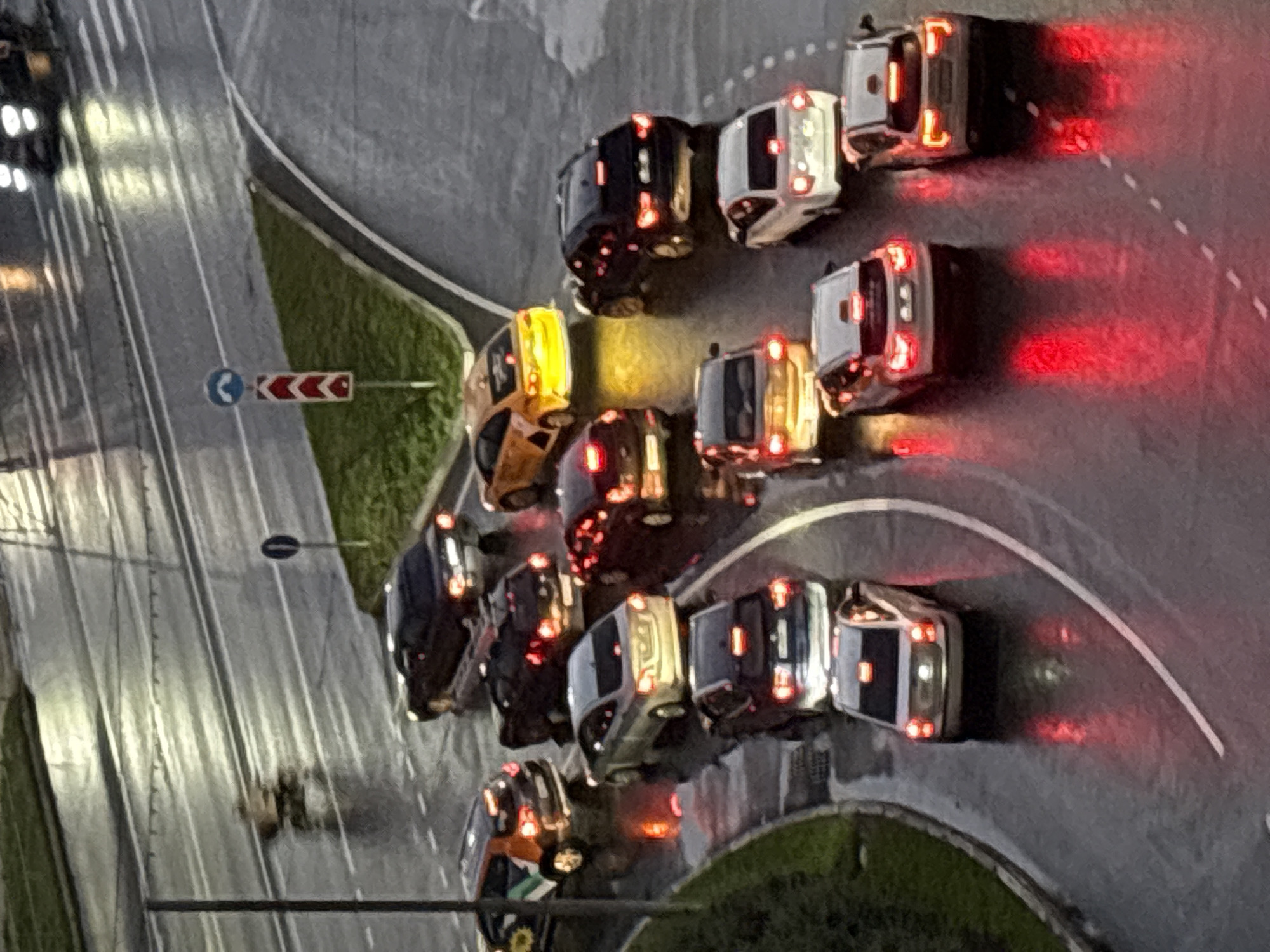 This aerial-view, low-light photograph captures a traffic jam on a wet, paved road, likely in Лесные Поляны, Russia. The scene is dominated by numerous cars, some with their brake lights illuminated, suggesting the vehicles are stationary or moving slowly. The road appears to be a multi-lane intersection or a stretch of highway with a curve.
The time of day is dusk or night, as indicated by the low illumination and the glowing lights of the vehicles. The wet surface of the road reflects the lights, enhancing the moody atmosphere. A patch of green grass is visible to the left of the road, with a traffic sign featuring a blue circle with a white arrow and a red and white directional arrow sign to its right, which suggests a turn or lane guidance.
The cars are a mix of colors and models, but their details are somewhat obscured by the lighting conditions and the perspective. There are no discernible people visible in the cars or on the road. The overall impression is one of stalled progress and a somber, perhaps frustrating, moment for the drivers caught in the congestion. The weather appears to be damp, contributing to the reflections on the road surface.