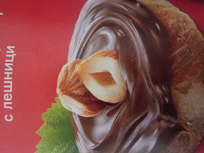 This image presents a close-up, two-dimensional graphic display, originating from the city of Balgarska Poliana, Bulgaria. The central subject is a round slice of light brown, textured bread or pastry, horizontally positioned. This base is generously covered with a thick, glossy, dark brown spread, which exhibits subtle concentric swirls and peaks, indicative of a viscous chocolate or hazelnut product. Two shelled hazelnuts are placed on top of the spread: one is whole, and the other is bisected longitudinally, revealing its lighter inner kernel. A partial green leaf, featuring serrated edges, is visible in the upper left portion of the image, positioned behind the hazelnuts. The background is a solid, vibrant red color. White Cyrillic text, partially visible at the top, reads "с лешници," which translates to "with hazelnuts." No human figures, actions, or direct interactions are depicted. The overall visual suggests a promotional photograph or a segment of product packaging.