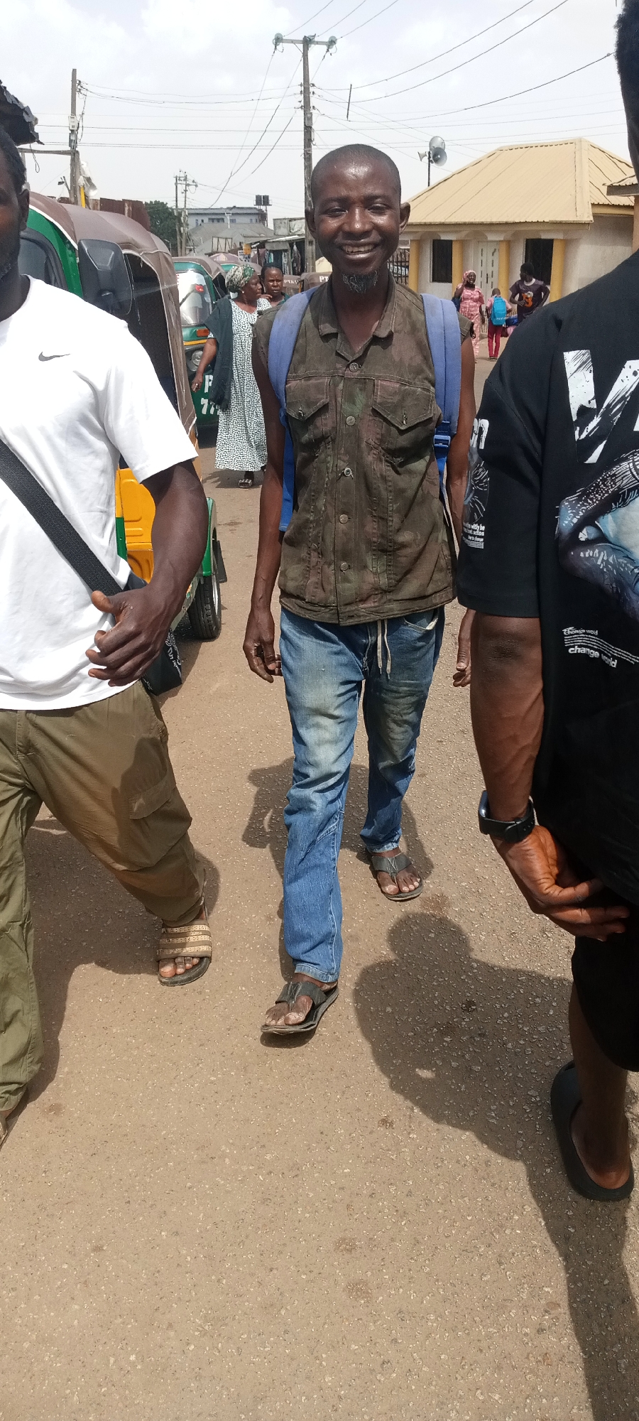 The image captures a bustling street scene in Jos, Nigeria, under bright, sunny daylight, indicated by strong shadows on the ground.
The central figure is a bald man with a short, dark goatee, wearing a camouflage-patterned sleeveless button-up shirt, blue jeans with a white drawstring, and dark sandals. He carries a blue backpack and is smiling broadly towards the camera, conveying a friendly or happy demeanor.
To his left, partially visible, is a man wearing a white t-shirt with a black Nike logo on the chest, olive-green cargo pants, and striped sandals. A black sling bag is draped across his body.
To the right, also partially visible, is a man wearing a black t-shirt with white graphic designs; text partially visible on his shirt includes "change world". He wears a black watch on his left wrist and dark slippers.
In the background, the street is active with several other people walking, including women in patterned clothing and children. Multiple motor tricycles, commonly called Keke Napep in Nigeria, are visible; one green and brown tricycle has the partial text "P 776" on its side. Buildings with varied architecture, including a light-colored structure with a prominent yellowish-orange roof, line the street. Overhead, utility poles and a network of power lines are visible against the bright sky. The scene depicts a vibrant urban or semi-urban environment with pedestrian and vehicular traffic.