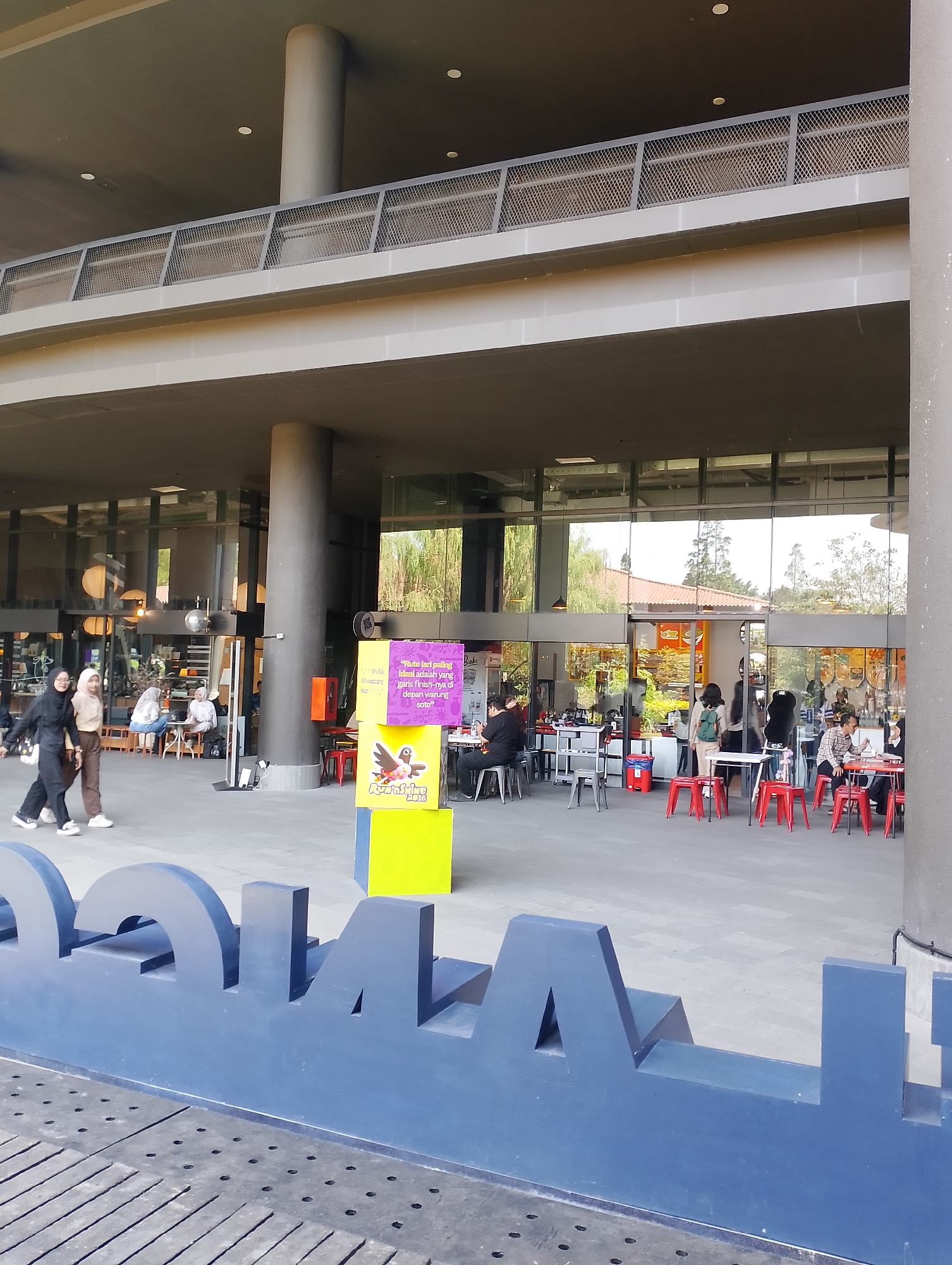 The image shows an outdoor seating area in front of a modern building with large glass windows, likely a cafe or restaurant, in Depok, Indonesia. In the foreground, a large blue art installation with cutout letters is visible. The ground is paved with grey tiles.
Several people are present in the scene. Three women, two in black and one in a beige hijab, are walking from left to right in the middle ground. Inside the building and in the outdoor seating area, other individuals are seated at tables. One group of three people, two women and one person of indeterminate gender, are sitting at a table inside, with one woman appearing to be talking. In the outdoor seating area, several tables with red chairs are occupied by diners.
To the right of the image, three colorful cubes are stacked, with the top cube featuring text and a graphic of a bird. A large dark grey column is to the right of these cubes. The building's facade features a dark grey overhang and extensive glass windows reflecting the outdoor environment. Trees and a red-tiled roof are visible in the reflections. The overall atmosphere suggests a casual, public gathering space.