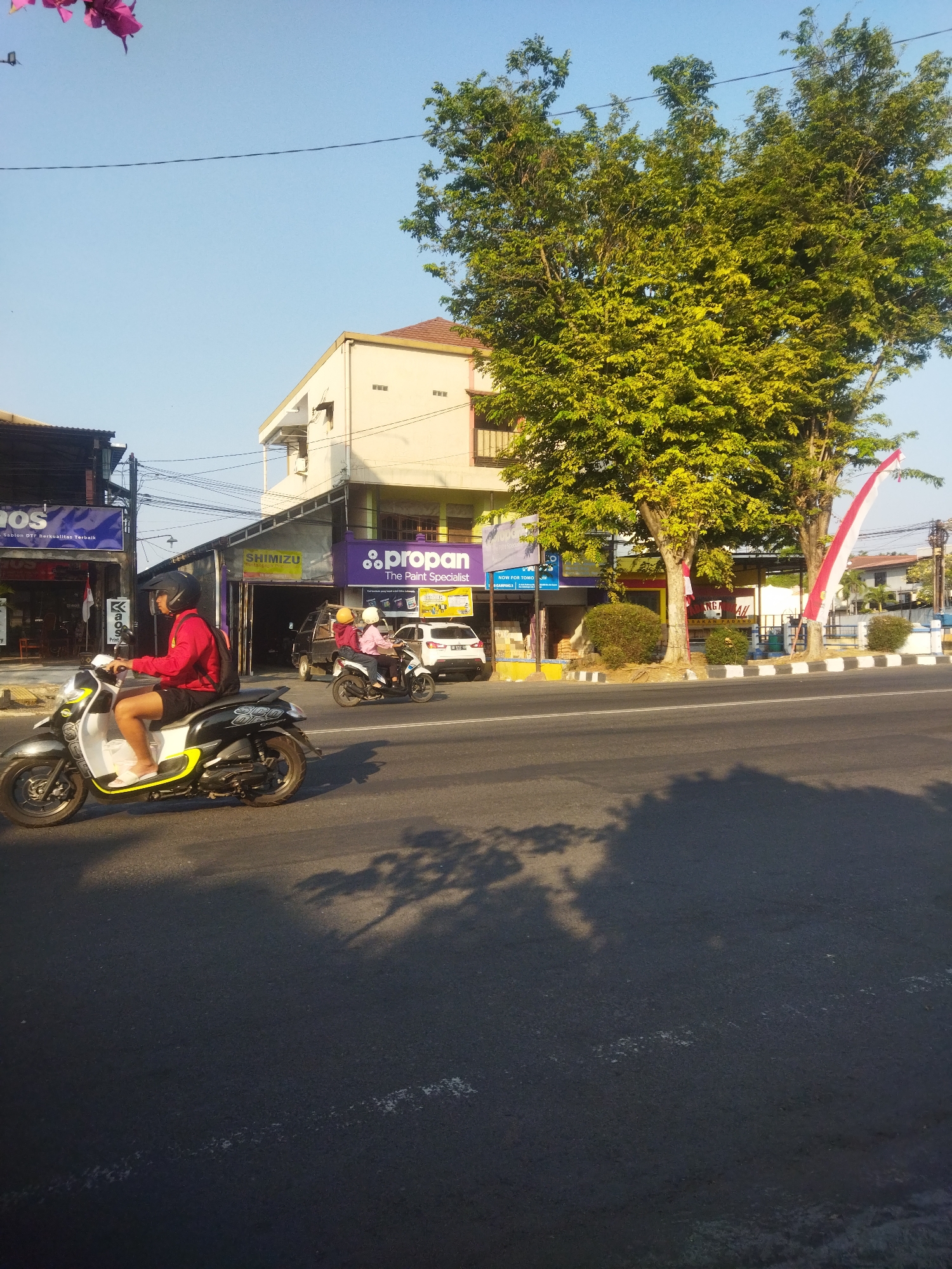 The image depicts a street scene in Sukoharjo, Indonesia, under a clear, pale blue sky. In the foreground, an asphalt road is visible with shadows cast from trees. A motorcycle, predominantly black and white with yellow accents, is being ridden by a person wearing a red jacket and black shorts, a helmet, and a backpack. The motorcycle has the branding "SCOOPY" on its side. Further down the road, another motorcycle carries two individuals, one in pink and the other in yellow, also wearing helmets. A white car is parked on the side of the road.

Along the street, buildings house various businesses. One building has a prominent purple sign that reads "Propan The Paint Specialist." Another sign displays "SHIMIZU." A building with a white facade and brown trim features multiple windows. Trees with dense green foliage line the street, and two Indonesian flags, red and white, are attached to poles. The environment suggests a commercial area in a city, with overhead wires and a sidewalk visible. The lighting indicates it is daytime, likely late afternoon or early morning due to the long shadows.