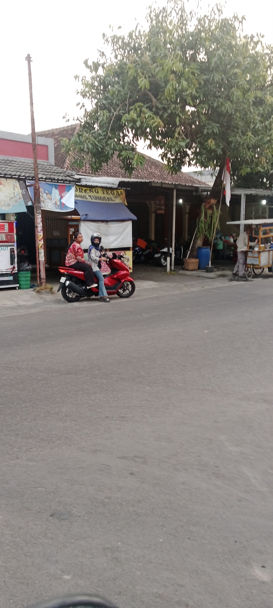 In Maguwoharjo, Indonesia, a person wearing a helmet and a patterned shirt is riding a red scooter. A second person, also wearing a helmet and a patterned top, is a passenger on the scooter. They are traveling on a paved road in front of a low-rise building with a red and gray facade and a tiled roof. A large tree with green leaves is situated to the right of the building, partially obscuring a red and white Indonesian flag attached to its trunk. A vendor's cart with a white canopy is visible further down the street, and several other scooters are parked nearby. The sky is overcast.