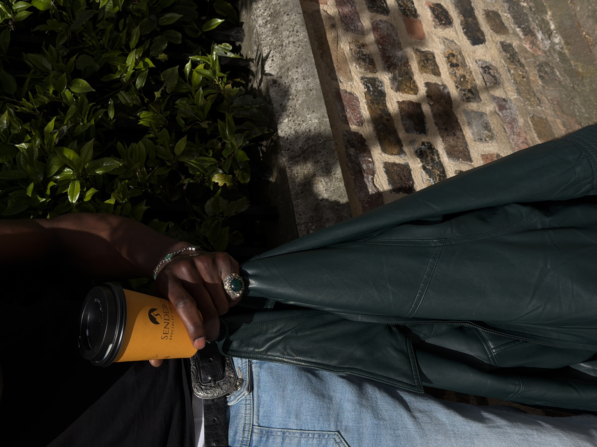 A medium shot shows a person holding a to-go coffee cup, with a dark green leather jacket draped over light blue denim jeans. The person has a silver bracelet and a large silver ring with a green stone. A black belt with a silver ornate buckle is visible. The person is wearing a black shirt. The background includes a brick wall and green leafy bushes. The location is suggested as a city street, possibly in London, United Kingdom.