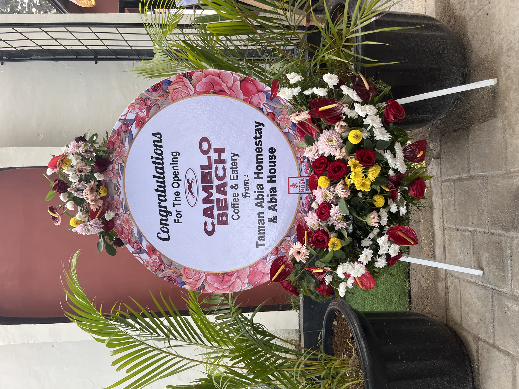 A floral arrangement sits on a stand in front of potted plants and a building. The floral arrangement is circular with a white center and a decorative border of pink and red flowers. Text on the white center reads "Congratulations For The Opening CAMELO BEACH Coffee & Eatery From: Taman Abia Homestay & Abia House". A small logo is visible at the bottom of the arrangement. The building behind the arrangement is light-colored with a darker section and a barred window. Potted palm plants are on either side of the arrangement. The ground is paved with interlocking bricks. The scene appears to be an outdoor setting in Kuta, Indonesia.