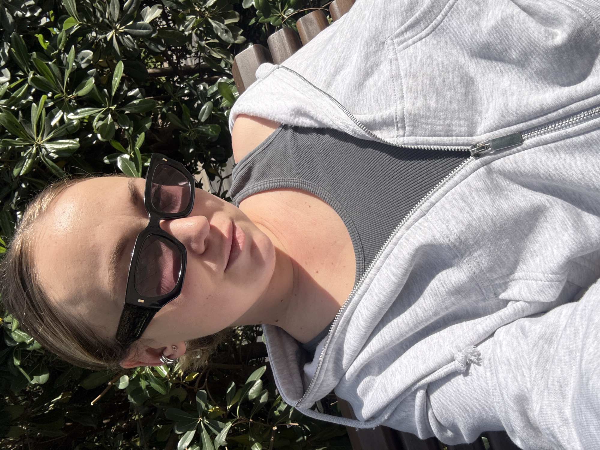 A woman is reclining outdoors on a wooden bench. She wears black sunglasses, a gray top, and a partially-zipped light gray hoodie. Lush green vegetation surrounds the bench. The location is the city of Bar, Montenegro.