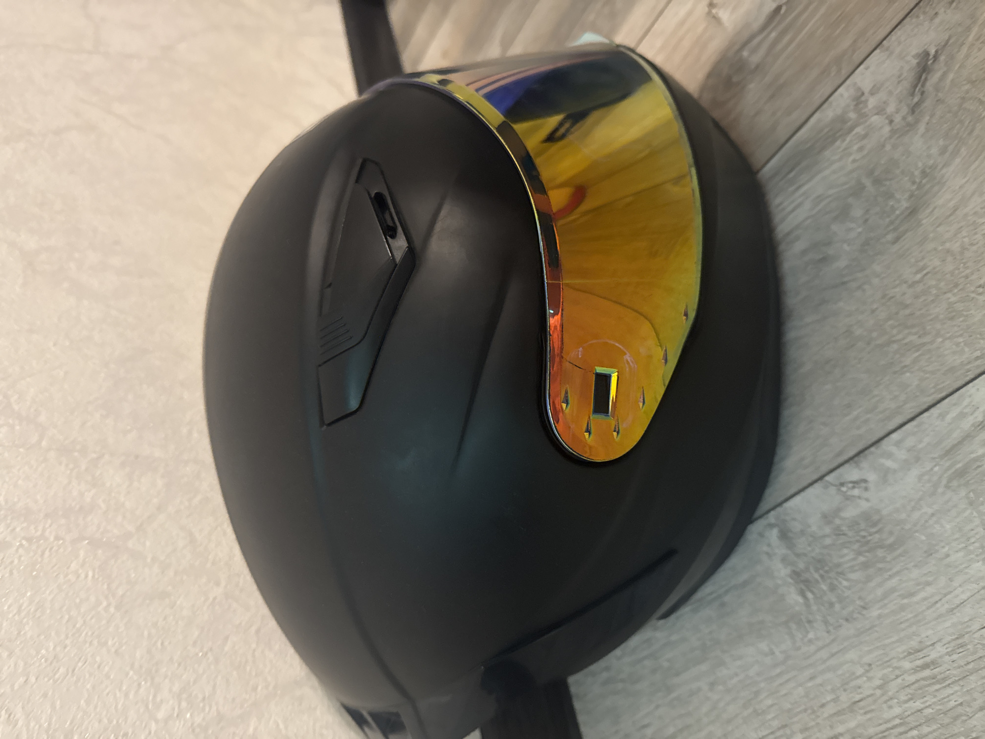 A matte black motorcycle helmet with a reflective gold visor rests on a light gray wooden floor. The helmet is positioned at an angle, with the visor facing the viewer and reflecting a colorful scene. The background consists of a textured, light-colored wall.