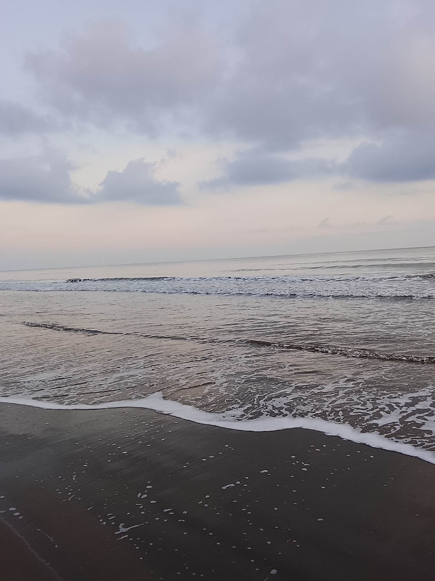 A serene beach scene at dusk in Reudeuep, Indonesia, is depicted. The image captures the gentle ebb and flow of the ocean against a dark, wet sand shore. The sky above is overcast with soft, diffused clouds, hinting at the approaching evening or a transition in weather. The water is calm, with small waves lapping at the shore, creating foamy white patterns on the sand. The overall atmosphere is one of tranquility and natural beauty, characteristic of coastal areas in Indonesia.