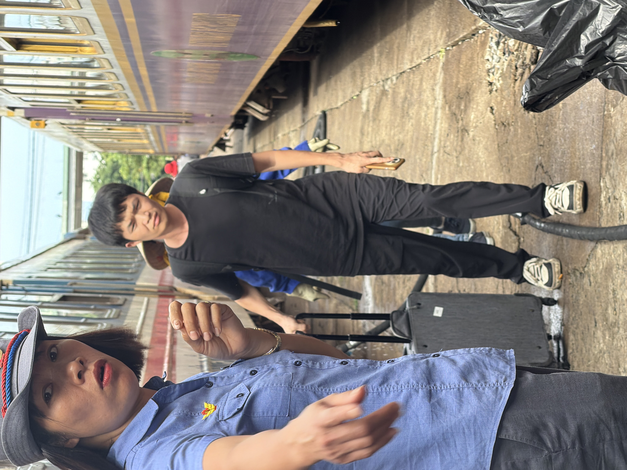 A group of people, including a woman in a blue collared shirt and a grey hat, a man in a black t-shirt and black pants, and others, are standing on a concrete platform next to a train. The woman in the foreground appears to be talking or reacting to something, with her mouth open and eyes wide. She is wearing a blue collared shirt with a yellow and red emblem and a grey hat with a red, white, and blue band. She is also wearing a gold bracelet. The man behind her is looking forward with a neutral expression. He is wearing a black t-shirt, black pants, and white sneakers. He is holding a brown smartphone in his right hand. A rolling suitcase is visible on the ground near the woman, and a black plastic bag is in the lower right corner of the image. The environment appears to be an outdoor train station, with the train cars running parallel to the platform. The scene suggests a travel or transit context, potentially at the Đà Nẵng railway station, given the location information.