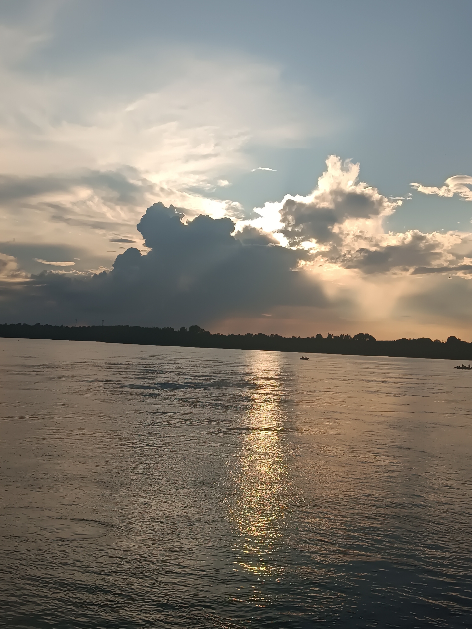 The image depicts a wide, calm river flowing in front of a silhouetted treeline under a dramatic sky. Sunlight streams through breaks in the clouds, casting golden rays onto the water's surface, creating a shimmering reflection. Two small boats with occupants are visible on the river, one in the mid-ground and another further in the distance. The scene captures a serene sunset over the Kama River, likely near Chaikovsky, Russia, a city known for its hydroelectric power station located on this river. The environment is natural and tranquil, with the focus on the interplay of light and water.