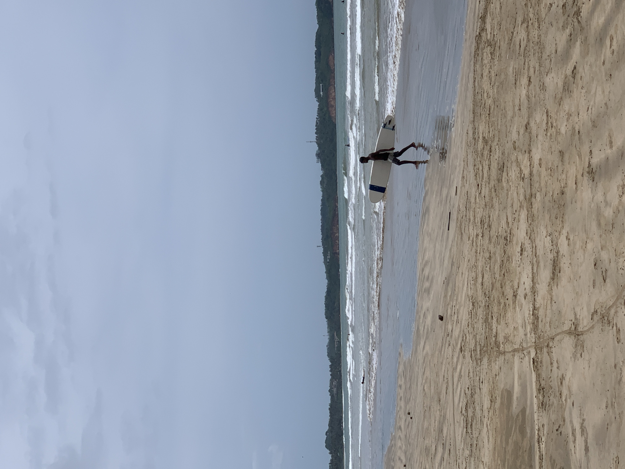 A wide shot depicts a beach scene in Weligama, Sri Lanka, under a cloudy sky. A person is walking along the edge of the water, carrying a surfboard. The beach consists of sand, and the ocean waves are visible. The shoreline is lined with vegetation.