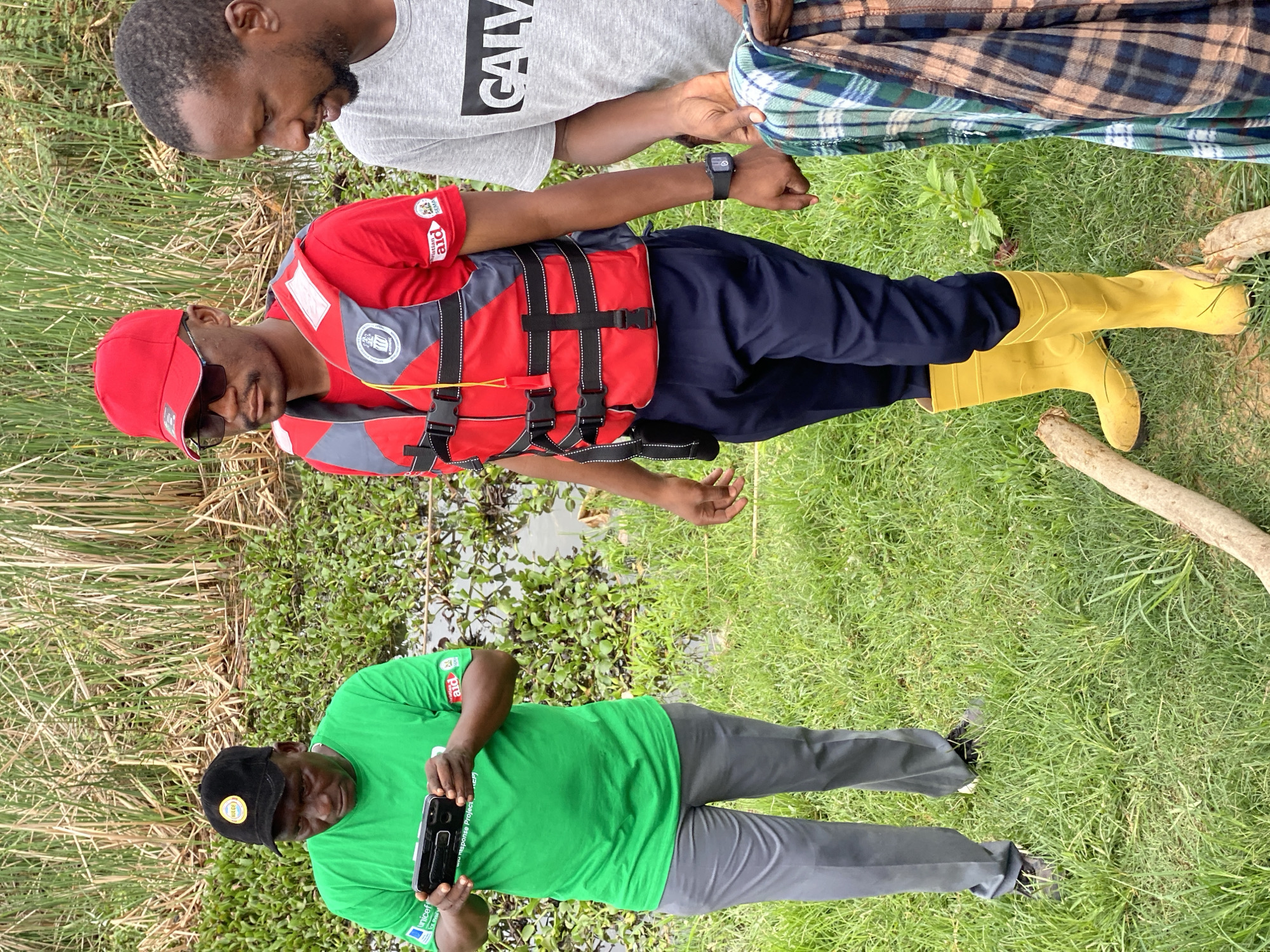 Three men are standing in a grassy area next to a body of water with dense vegetation. The man on the left is wearing a green t-shirt, gray pants, and a black baseball cap with a yellow logo. He is holding a black smartphone and appears to be looking at it. The man in the center is wearing a red t-shirt, navy blue pants, yellow rubber boots, a red baseball cap, and a red and gray life vest. He is looking forward. The man on the right is wearing a gray t-shirt and is holding a plaid blanket. He is looking down. The background consists of tall reeds and aquatic plants. The location is Kawo, Nigeria.