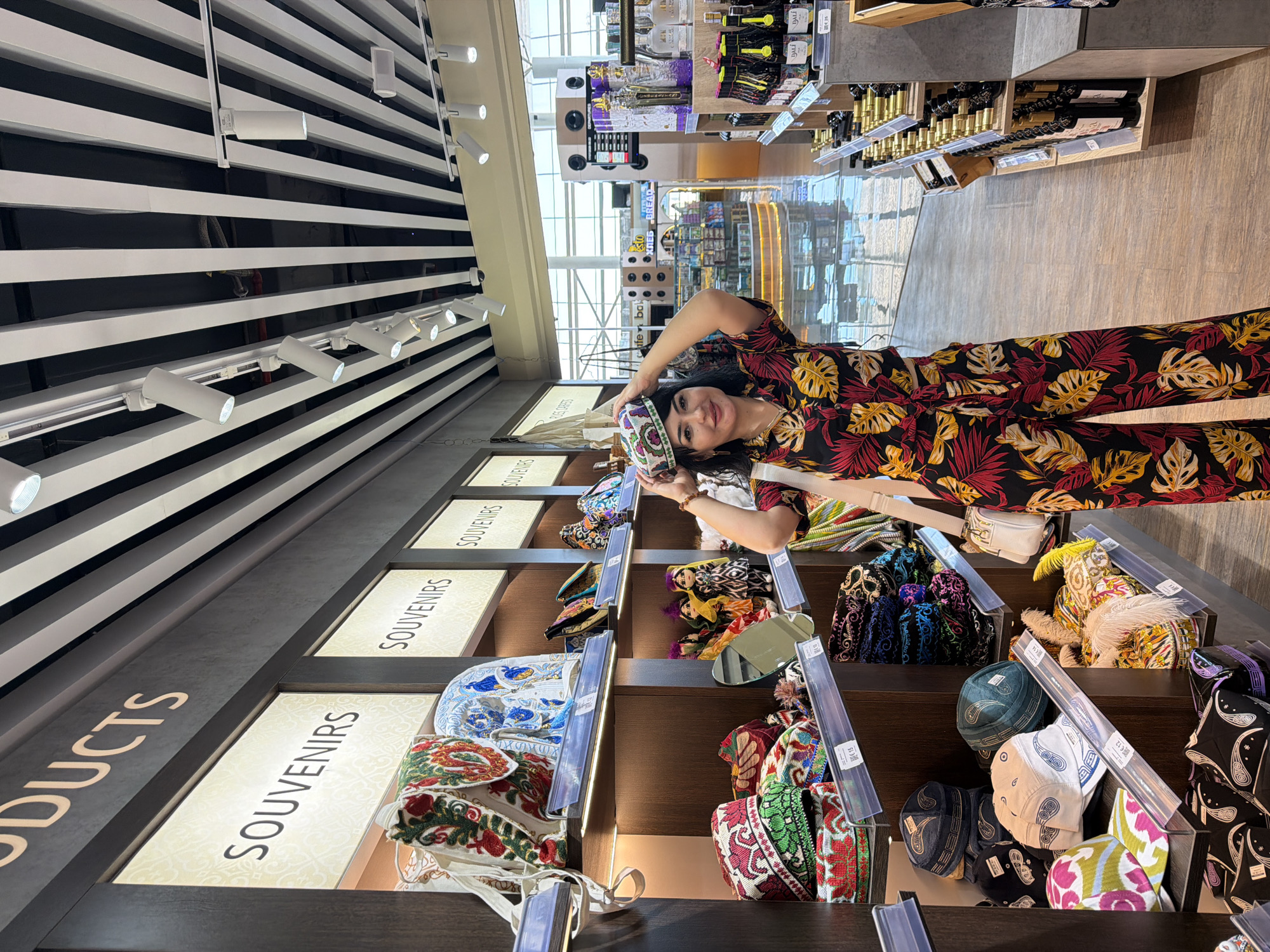 The image depicts a woman posing with a patterned hat on her head in a souvenir shop. She is wearing a brightly colored, tropical-print jumpsuit. The shop shelves are stocked with various souvenirs, including embroidered hats, bags, and dolls, displaying traditional Uzbek craftsmanship. The background suggests a modern airport or shopping mall with rows of products, including bottles of wine on a shelf to the right. The overall ambiance is that of a retail environment offering local crafts to travelers.