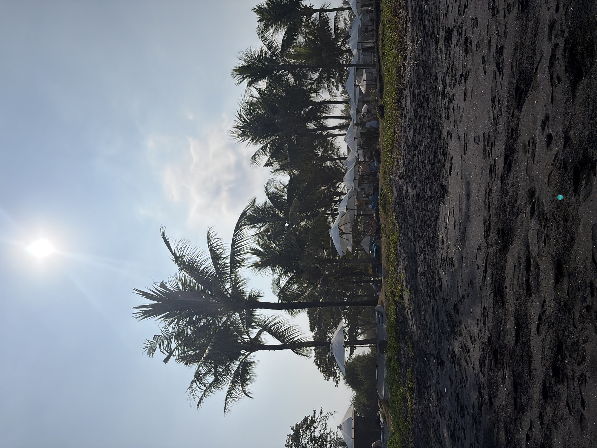 The image depicts a tropical beach scene in Blahbatuh, Indonesia, under a bright, hazy sun. The foreground is dominated by dark, coarse sand with numerous footprints, suggesting recent activity. In the midground, a line of white beach umbrellas is set up, providing shade to lounge areas. Behind the umbrellas, a dense line of palm trees stretches across the frame, their fronds silhouetted against the bright sky. A few people are visible in the midground, scattered among the umbrellas, some appearing to be lounging or walking. The vegetation behind the umbrellas is lush and green. The sky is a pale blue with some scattered, wispy clouds. The overall impression is of a relaxed, sun-drenched beach resort environment.
