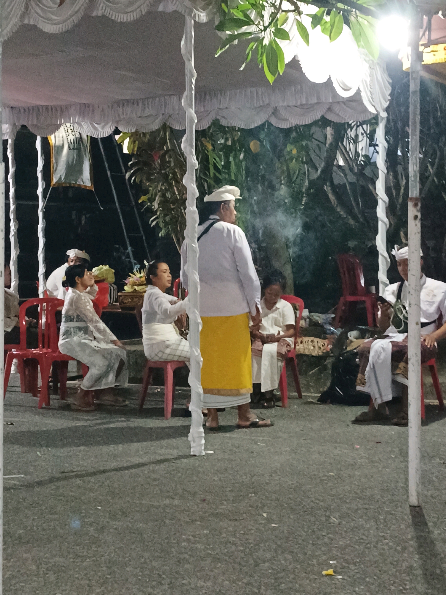 The image depicts an outdoor gathering under a white tent, likely in Kediri, Indonesia, given the presence of what appears to be Balinese traditional attire on several individuals. The scene is dimly lit, with artificial lighting casting shadows. In the foreground, a person stands facing away from the camera, wearing a white top and a yellow sarong. To the left, two women are seated on red chairs. The woman on the far left wears a light-colored, patterned dress, while the woman closer to the center is dressed in white with a white sash. To the right, another individual, also in white attire and a head covering, is seated on a chair, looking down at an object in their hands, possibly a phone. In the background, more people are visible, some seated, and there are indications of a ceremonial setup with offerings or decorations. The ground is asphalt, with scattered debris and shadows indicating the presence of artificial lights. The overall atmosphere suggests a cultural event or religious ceremony taking place at night.