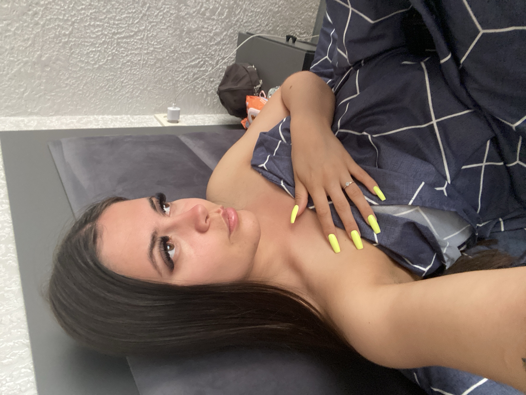 The image shows a young woman lying in bed, covered by a navy blue duvet with a white geometric pattern. She is looking to her right, with her left arm resting on her chest and her right hand on the duvet. She has long, dark brown hair, dark eyeliner, and bright yellow painted fingernails. A ring is visible on her left hand. The background shows a grey headboard and a textured white wall with a light switch and a power outlet. A dark brown baseball cap and an orange and white package are on a dark bedside table to the right. The image is a close-up shot, focusing on the woman and her immediate surroundings. There is no information in the image to suggest the location is the city of Chalda, Russia.