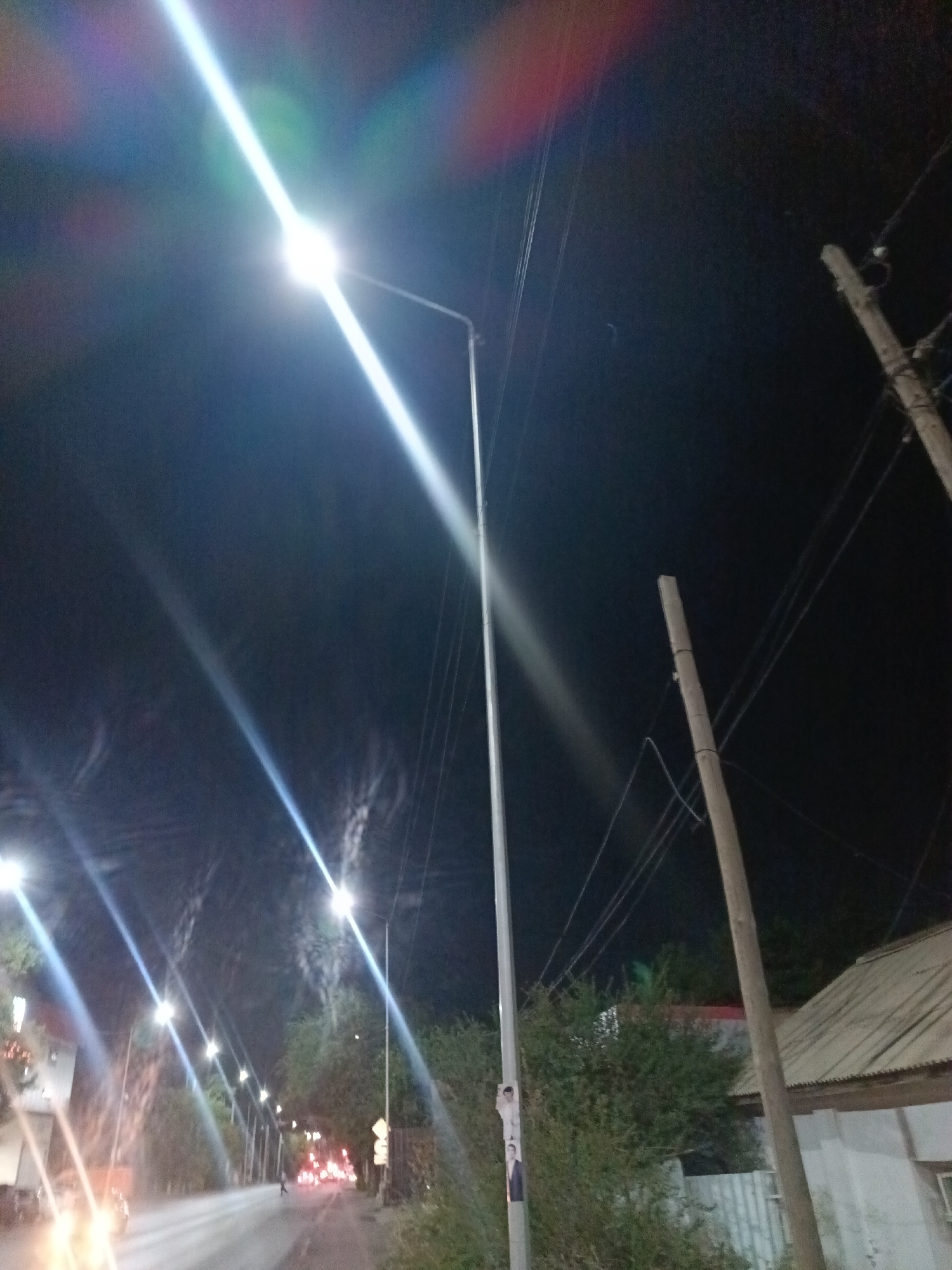 This is a nighttime photograph taken on a street in Shymkent, Kazakhstan, likely in a residential or suburban area. The image is dominated by the bright glare of streetlights, which create strong light streaks and lens flares. The street appears to be paved, with vehicles visible in the distance, their headlights and taillights creating red and white glows. There are numerous utility poles and power lines crisscrossing the sky, and trees and bushes line the sides of the road. On the right side of the image, a building with a corrugated metal roof is partially visible, along with a wooden pole adorned with posters. The overall impression is of a dimly lit urban environment, illuminated by artificial lighting.