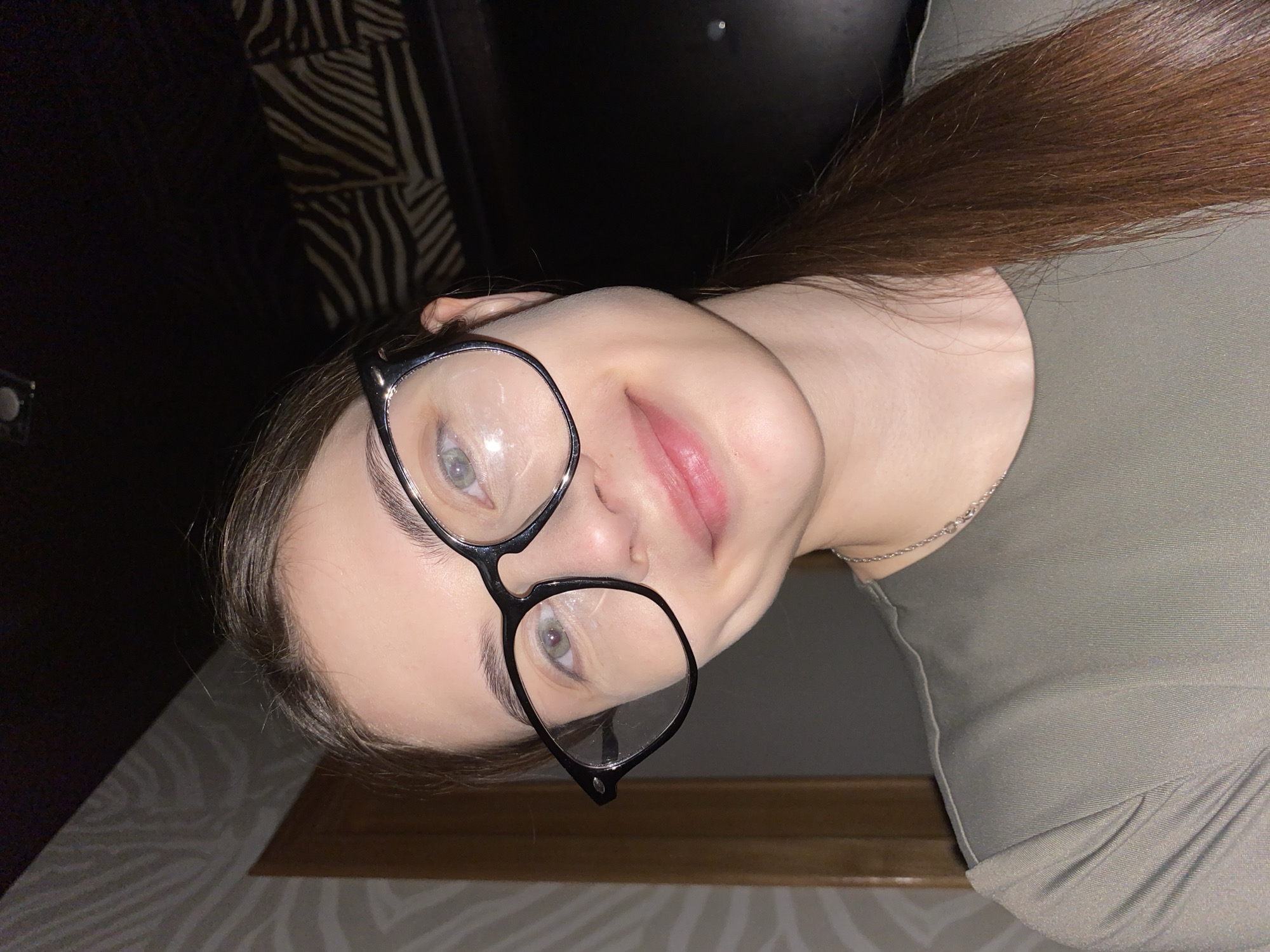 A close-up portrait features a young woman with a gentle, slightly smiling expression, her head tilted as she looks directly at the camera. She has long, straight reddish-brown hair and fair skin. She wears black-framed glasses, which reflect light and partially obscure her light-colored eyes. A thin, silver-toned chain is visible around her neck. She is dressed in an olive-green garment, possibly a top or dress, made of a textured or ribbed fabric.
The background is dark, indicating an indoor setting with artificial lighting, likely taken in the evening or at night. Behind her, a wall features a patterned design, possibly an abstract stripe or zebra print. A wooden element is also visible in the lower part of the frame. The overall mood is calm and friendly. No text is visible in the image. The location context for this image is Votkinsk, Russia.