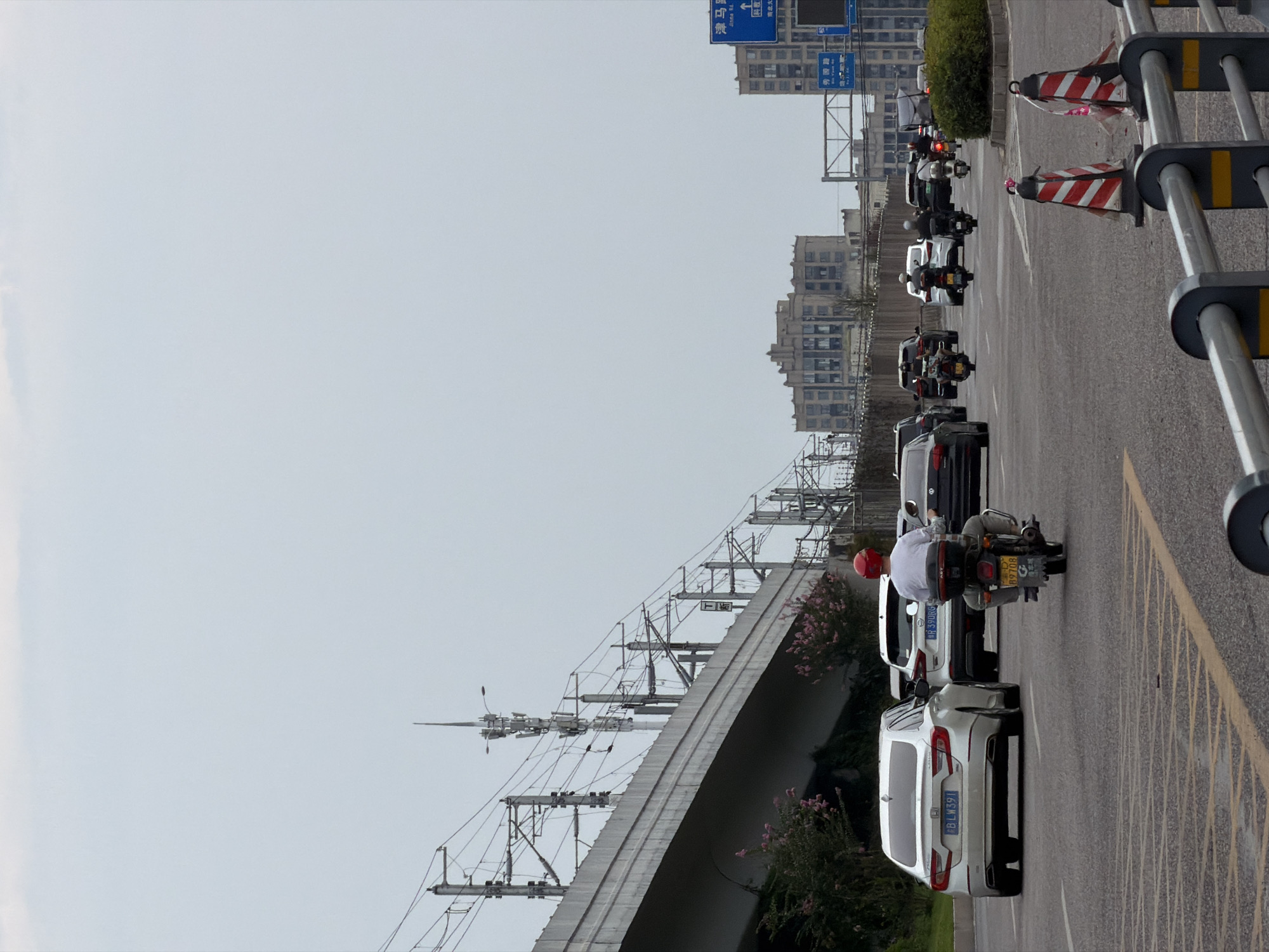 The image captures a street scene in Jiangjin, China, under an overcast sky. A multi-lane road dominates the foreground, with traffic flow from left to right. A white Honda sedan with license plate BLW391 is parked in the leftmost lane. To its right, a white SUV with license plate R39DRG is also present. A person wearing a red helmet and a white shirt is riding a motorcycle in the center of the road, heading towards the right. Several other motorcycles and cars are visible in the midground and background, indicating moderate traffic. An elevated structure with overhead railway lines runs along the left side of the image, with buildings visible in the distance. Traffic cones and a metal guardrail are present in the foreground. The overall atmosphere is that of a busy urban environment.
