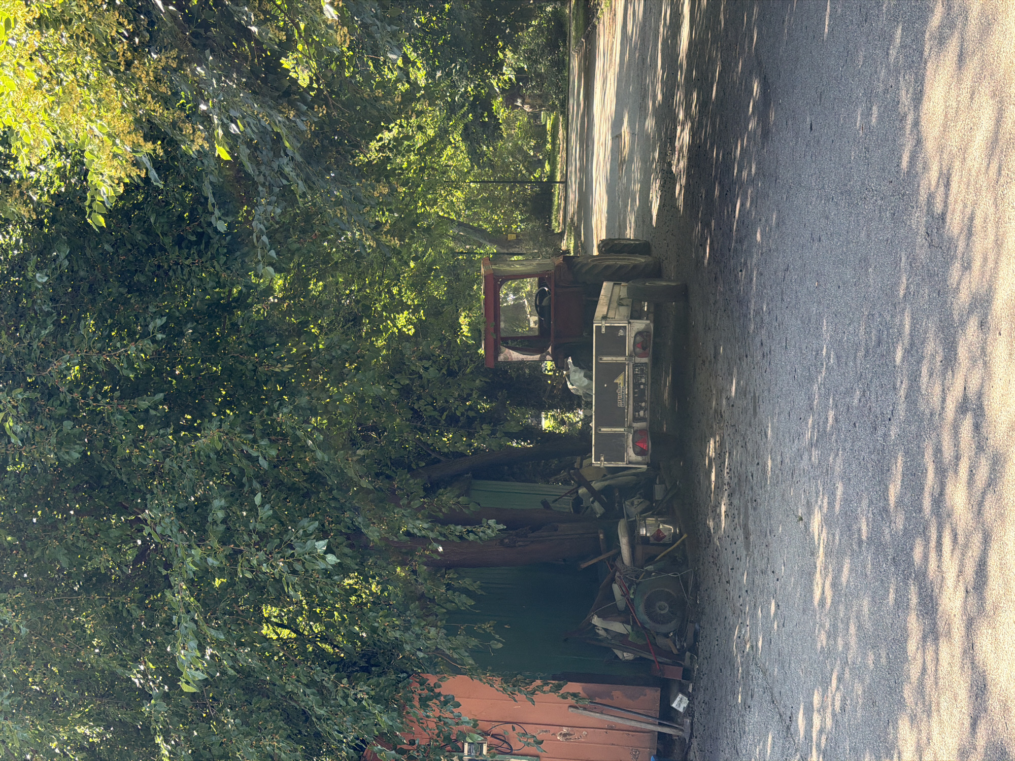 The image depicts an outdoor scene under the dappled sunlight filtering through a dense canopy of trees. In the foreground, a paved road is visible, with patches of gravel and debris scattered across its surface. The road is partially covered by the shadows cast by the foliage above.
To the left of the road, a collection of items are piled near a weathered, orange-colored structure and a green wall. These items appear to be discarded tools and equipment, including what looks like a wheel from a vehicle, various metal rods and pipes, and other miscellaneous objects.
Further into the scene, a red tractor is parked. It is attached to a small, flatbed trailer. The tractor has large rear wheels and a visible steering wheel inside its cab. The trailer has reflective taillights and some lettering on its rear panel.
The background shows a continuation of the road, which appears to curve to the right and leads into a densely wooded area. The trees are tall and leafy, creating a shaded environment. A lamppost is visible in the mid-ground. The overall impression is one of a semi-industrial or storage area situated near a park or a tree-lined street. The location is identified as being in the city of Pazardzhik, Bulgaria.