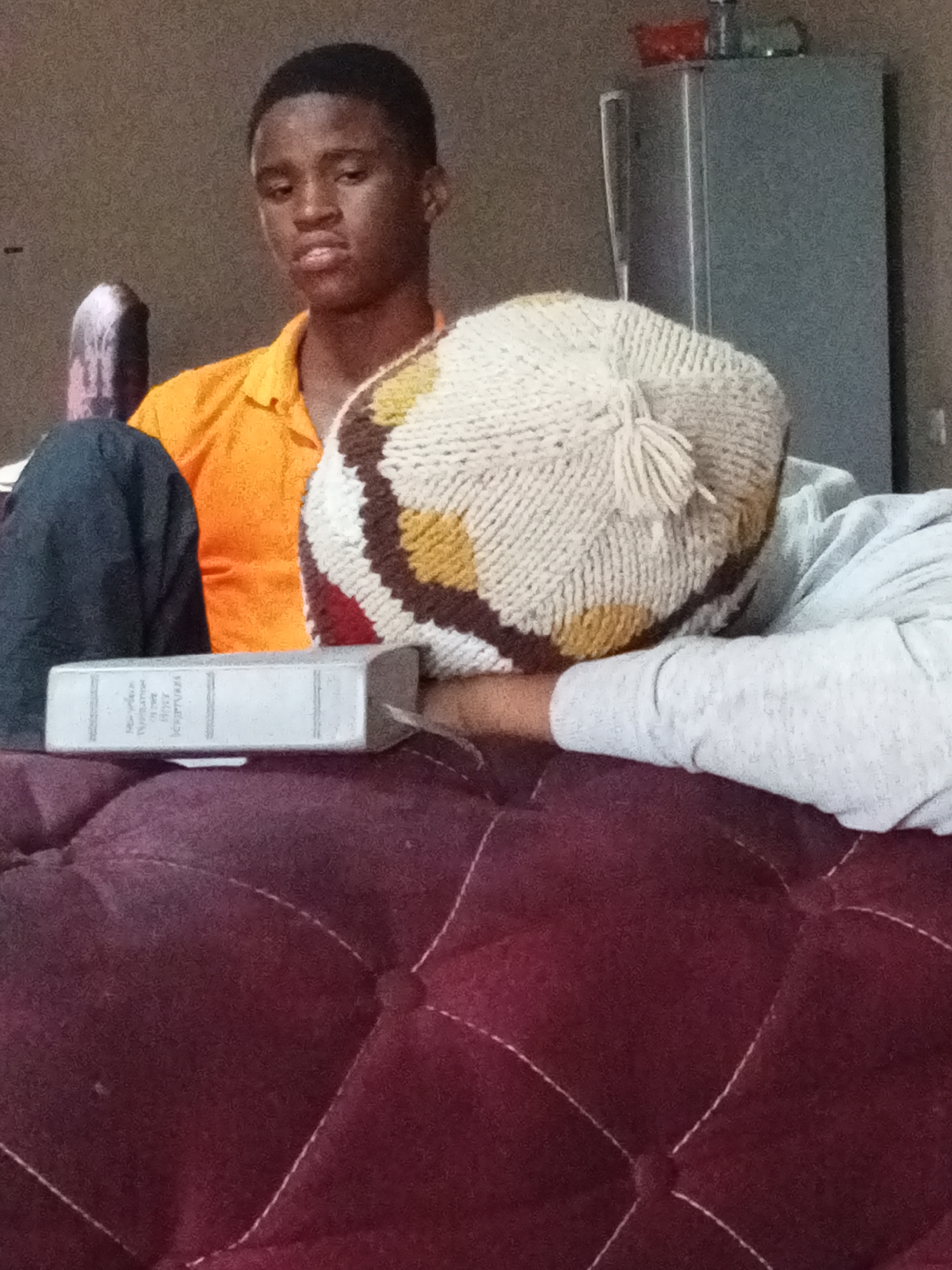 A young Black male wearing an orange polo shirt and a knitted cap is seated indoors, possibly in a living room in Jos, Nigeria. He is looking towards the camera with a neutral expression. In the foreground, a book with a silver cover lies on a plush maroon sofa. Another person, partially visible, is leaning on the sofa, wearing a light-colored hoodie and a similar knitted cap. A gray refrigerator is visible in the background, along with a wall and some items on top of the refrigerator.