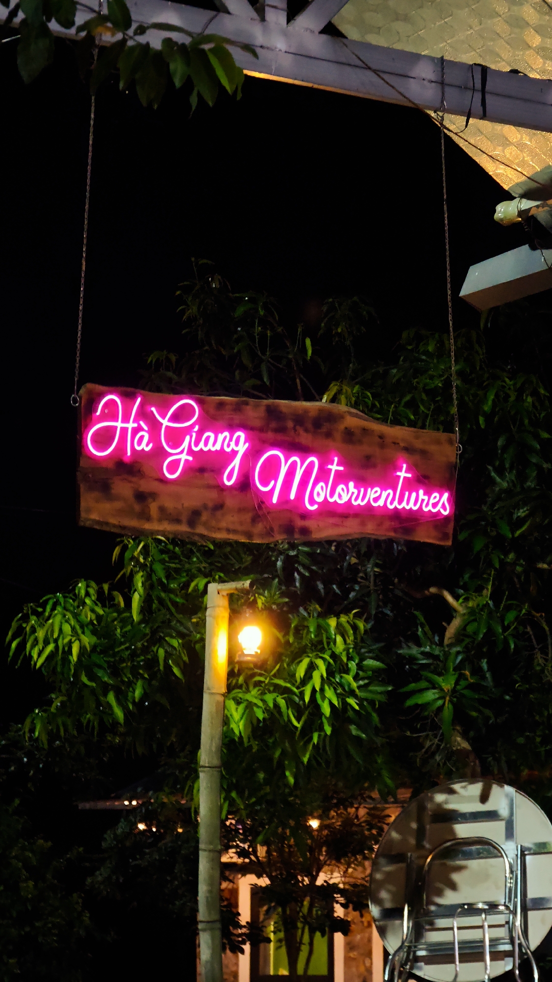 The image depicts a wooden sign with the words "Hà Giang Motorventures" written in bright pink neon lights. The sign hangs from a chain attached to a structure above. The background consists of dense green foliage and a lamp post with a glowing orange light. The image appears to be taken at night in Du Già Commune, Vietnam. A round, metal object is visible in the lower right corner of the image, possibly part of a vehicle.