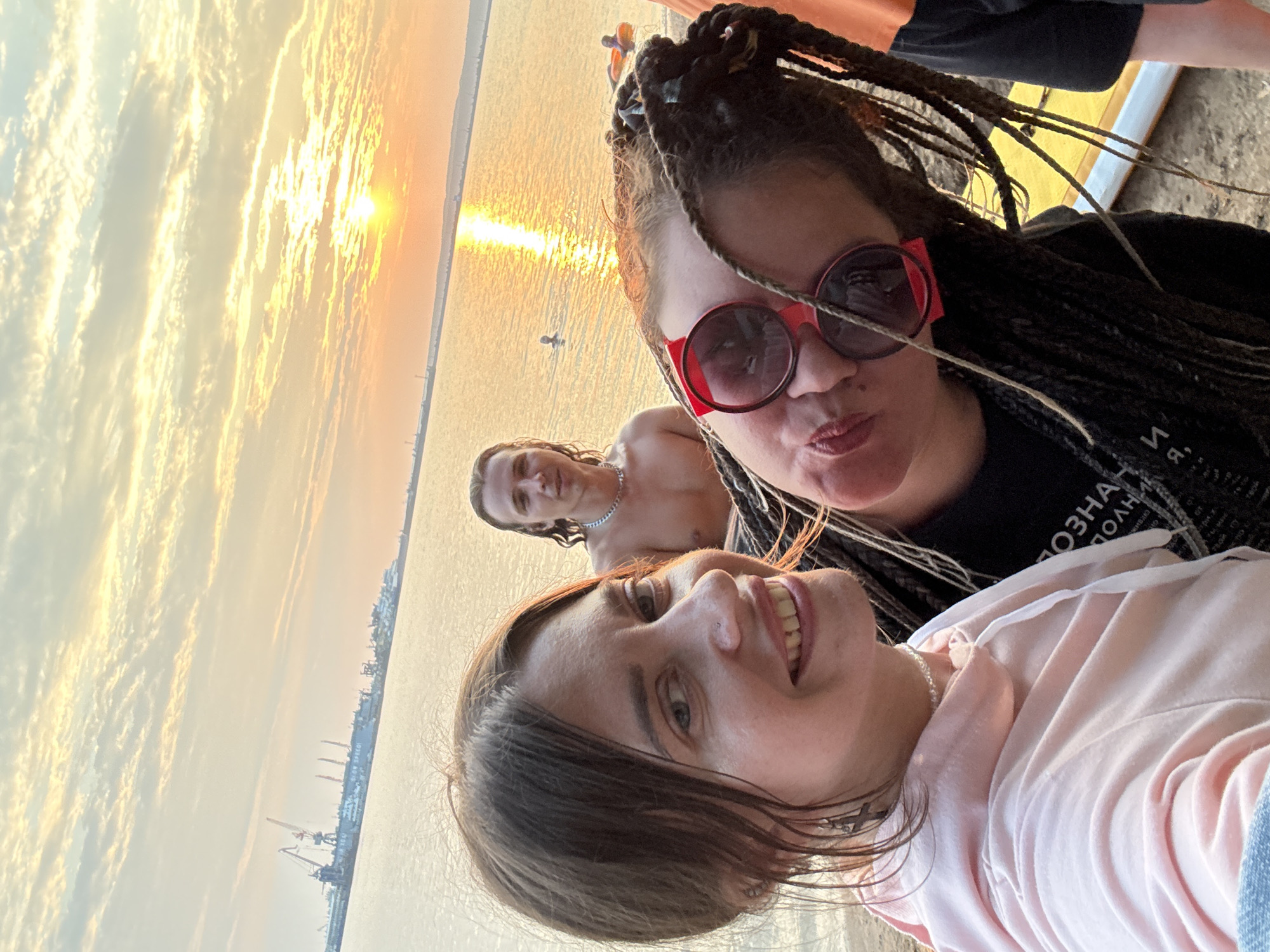 In a frontal medium shot, three individuals are positioned by a body of water during sunset. The woman in the foreground on the left smiles and looks at the camera, wearing a pink hoodie and a pearl necklace. To her right and slightly behind her, another woman is captured in a playful pose, leaning forward with her head tilted and wearing large red sunglasses. Her dark hair is styled in braids. Behind them, a shirtless man with long hair and a chain necklace looks toward the camera. The background features a calm body of water reflecting the golden hues of the sunset. In the distance, a shoreline is visible with industrial cranes and buildings. Two other individuals are present in the water, one with an inflatable tube. The overall scene depicts a casual outdoor gathering at dusk.