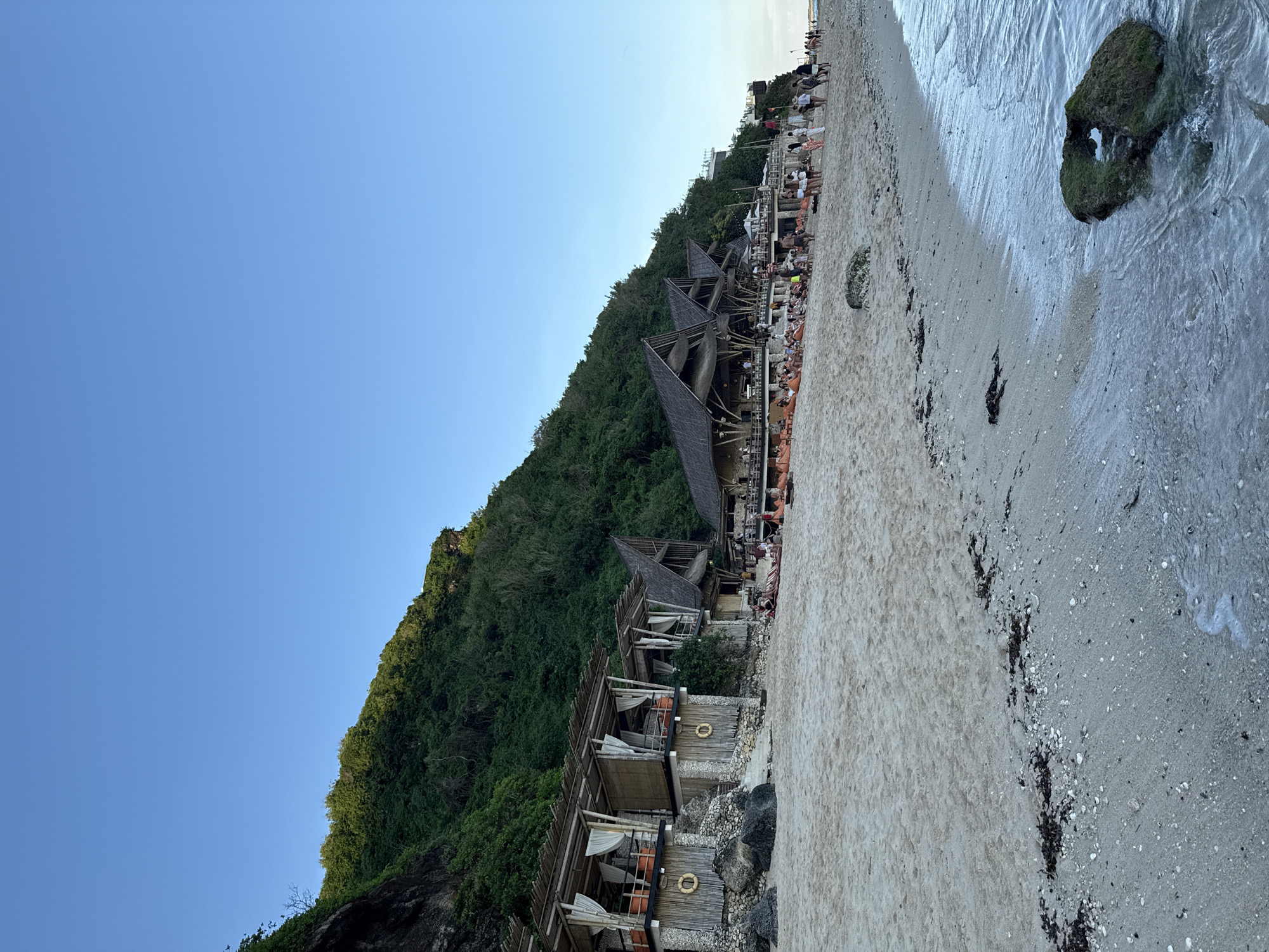 This is a medium shot of a beach in Ungasan, Indonesia. The image is taken from a low angle and features a clear blue sky filling the top left portion. A steep, green hillside covered in dense vegetation rises from the left edge, blending into the beach area. Along the hillside are several structures, including small beach bungalows with wooden frames, life preservers, and white curtains. Adjacent to these are larger open-air buildings with thatched roofs, appearing to be restaurants or bars. Numerous people are seated in these establishments. The beach occupies the right half of the image and is composed of sand with dark seaweed washed ashore. The ocean, visible on the far right, features a moss-covered rock protruding from the water. The scene appears to be set in the late afternoon or early evening, indicated by the soft light.