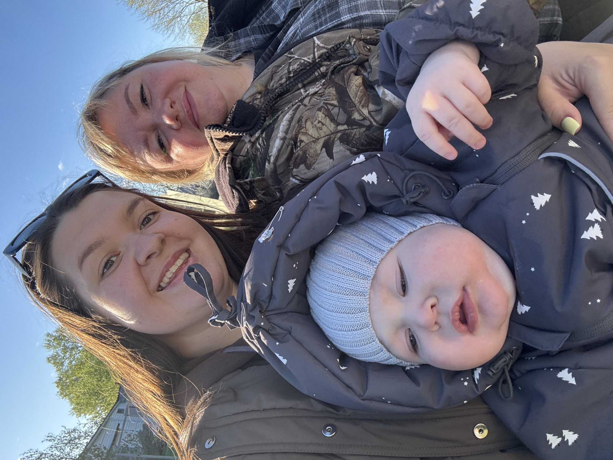 The image shows a selfie of two women and a baby. One woman is in the lower left, wearing sunglasses atop her head and a brown jacket. Another woman is in the upper right, wearing a camouflage jacket and a plaid shirt. The baby is wearing a grey hat and a grey snowsuit with white tree patterns. The background is blue sky and some greenery, likely trees. A small structure, possibly a shed, can be seen. The location of Lukintsy, Russia is not verifiable from the image. The scene appears to be taken outdoors in daylight.