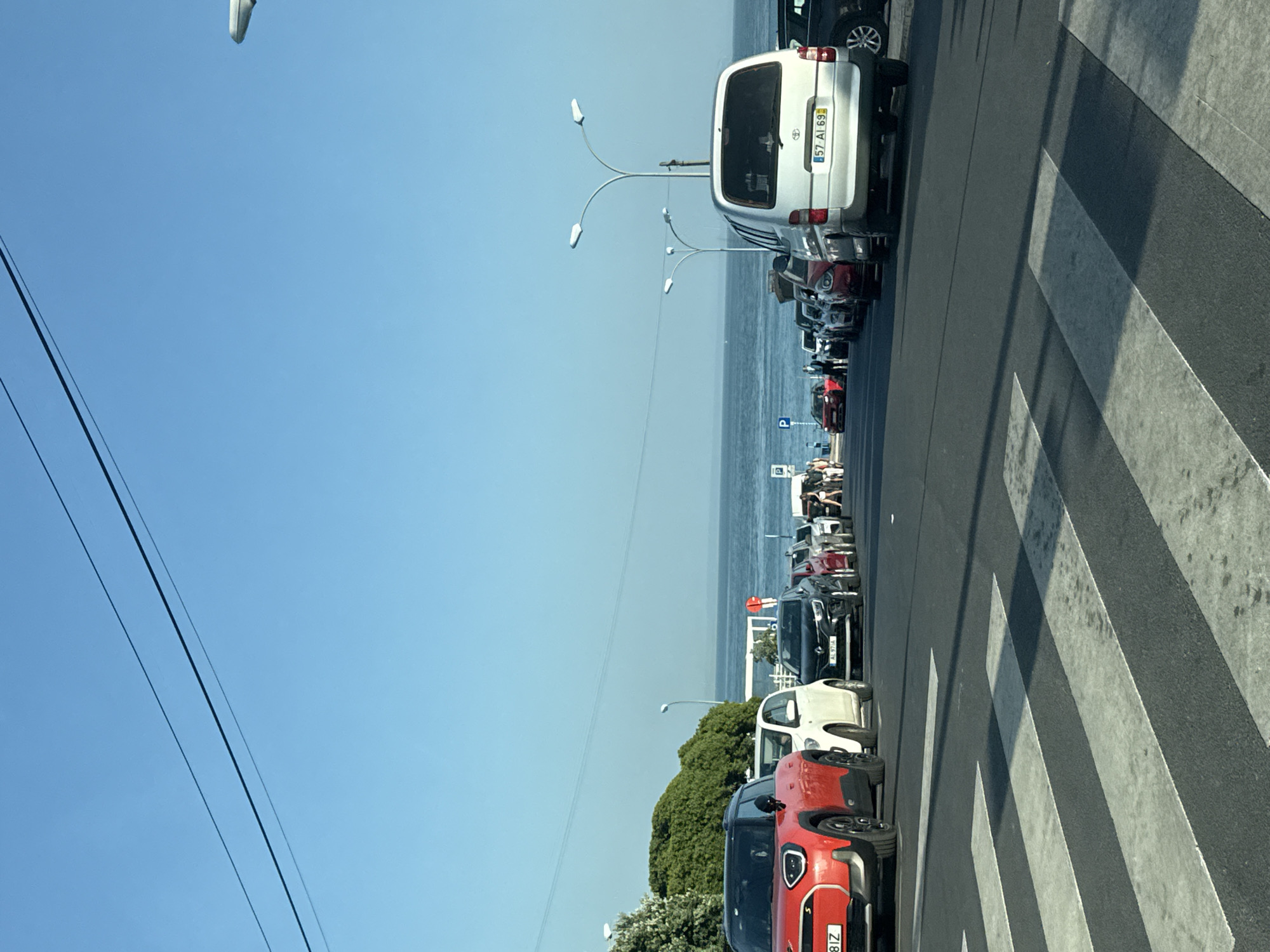 The image captures a street scene in Oeiras, Portugal, with a clear blue sky dominating the upper portion. The foreground features a black asphalt road with a prominent pedestrian crossing marked by white stripes. A line of parked cars is visible along the right side of the road, extending into the distance. The cars are of various colors and models, including a red Mini Cooper in the foreground on the left, a white car, and a silver van with a license plate reading "57 AI 69". Further down the street, more vehicles and a few pedestrians can be observed. Beyond the cars and road, a body of water, likely the Atlantic Ocean, stretches out, with a distant shoreline and some buildings visible. Trees and greenery are present on the left side of the image, bordering the road. Streetlights with multiple lamps are spaced along the roadside. The overall atmosphere suggests a sunny day in a coastal city.
