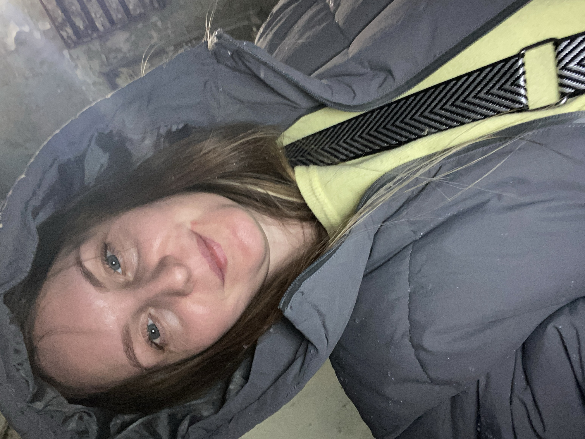 A woman with fair skin, light brown hair, and blue eyes is looking at the camera. She is wearing a gray puffy coat with the hood up and a yellow top with a black and white chevron-patterned strap across her chest. The background is slightly out of focus, with a texture suggesting a concrete or stone surface. The lighting appears to be dim. The setting is not reliably determinable to Noviy, Russia based on the visual information provided.