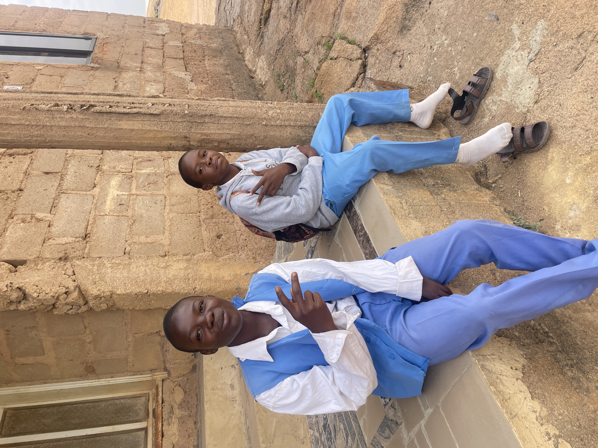 Two young boys are reclining on a stone structure in what appears to be Jos, Nigeria. The lower boy is wearing a blue and white uniform and makes a peace sign, while the upper boy wears a grey hoodie and blue trousers, also making a peace sign and wearing white socks with sandals. The structure is adjacent to a building with a window and a stone wall. The environment is arid.