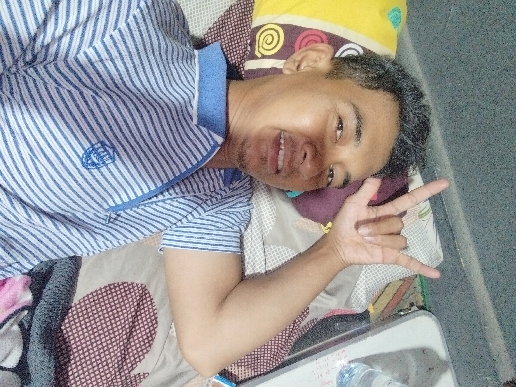 A man is lying on his back in Cemplang, Indonesia, wearing a blue and white striped shirt and making a peace sign with his left hand. He is smiling, and his eyes are open. The man is lying on a bed with striped pillows and a patterned blanket. A gray wall is visible in the background. A bottle of water is on a white surface in the lower right corner of the image.