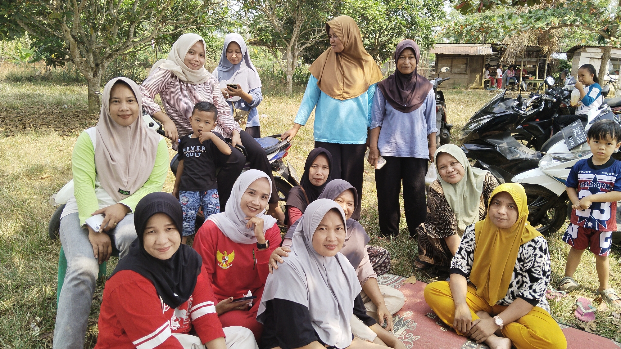 This outdoor, daytime photograph captures a gathering of approximately fifteen people, primarily women and children, in what appears to be a rural or semi-rural setting. The group is positioned in front of several parked motorcycles and some rudimentary structures, possibly stalls or small buildings. The environment is characterized by lush green grass and trees, suggesting a warm climate. The people are dressed in casual, modest attire, with many of the women wearing hijabs. Some individuals are seated on the ground, while others are standing or leaning. The overall atmosphere suggests a relaxed, communal event. The specific location is identified as Margatunggal, Indonesia.