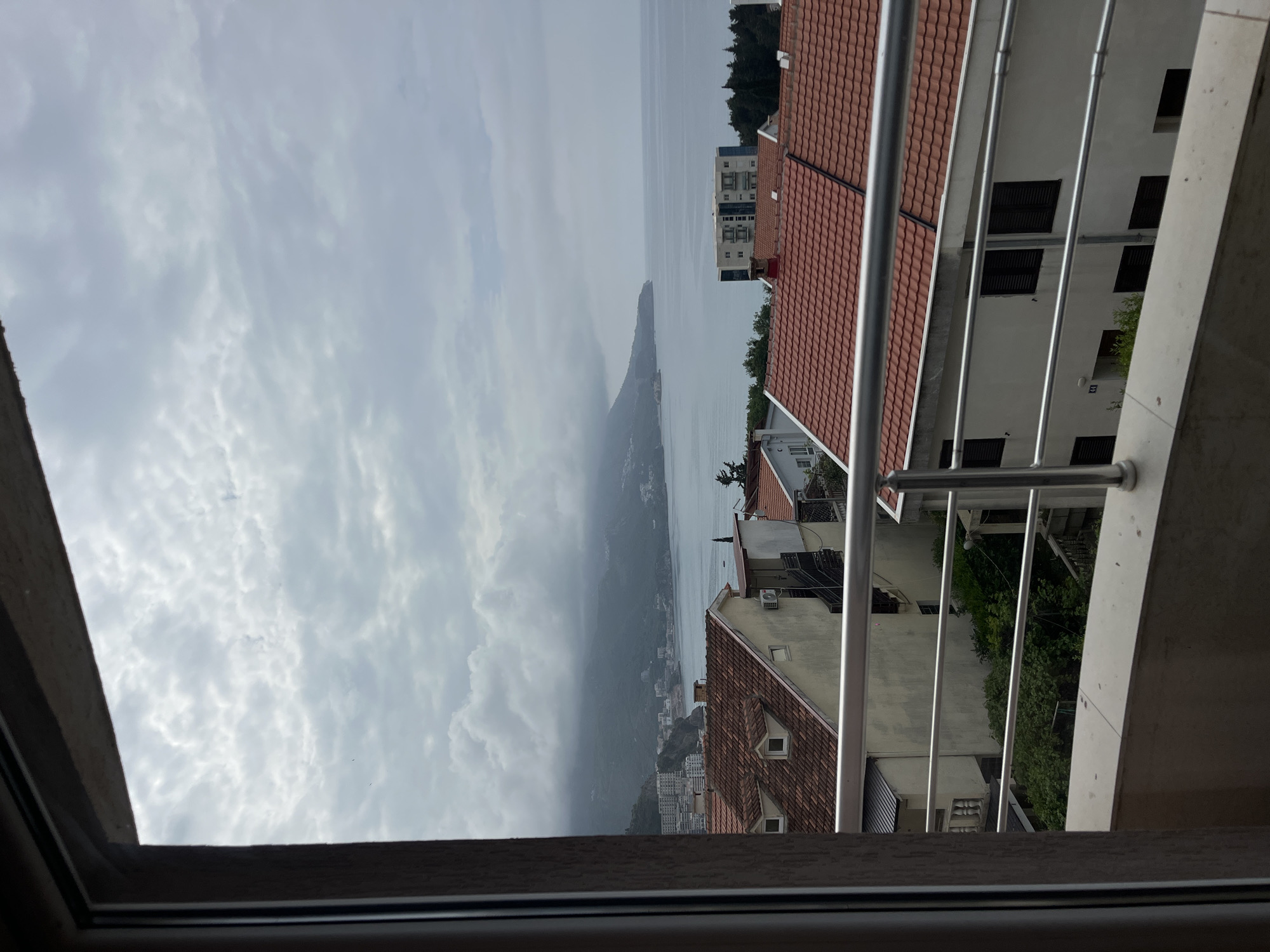The image shows a view from an elevated position, likely a balcony or window, in what appears to be a coastal city or town. The view includes residential buildings with red-tiled roofs. Further in the distance, there is a body of water with a mountainous landmass extending into it. The sky is overcast with heavy clouds. The window frame is visible at the bottom of the image. Boreti, Montenegro, is the suspected location. There are no people visible.