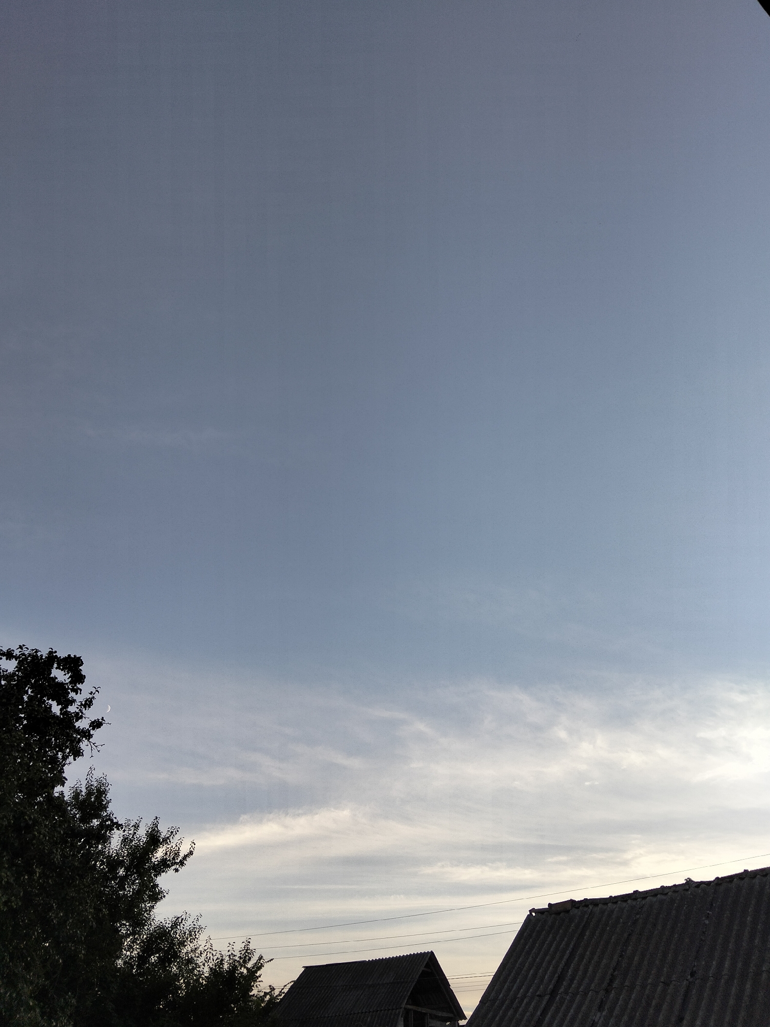 The image captures a view of the sky over Ustyvytsia, Ukraine. The sky is a pale blue with wispy white clouds. In the foreground, the dark silhouettes of trees with lush green leaves are visible on the left side of the frame. There is also a crescent moon visible in the upper left quadrant of the image, partially obscured by the tree. Two rooftops of houses, made of corrugated material, are present at the bottom of the image. One rooftop is angled and the other is partially visible on the right. Electrical wires are strung horizontally across the scene above the rooftops. The overall impression is a rural or suburban landscape at dusk or dawn.
