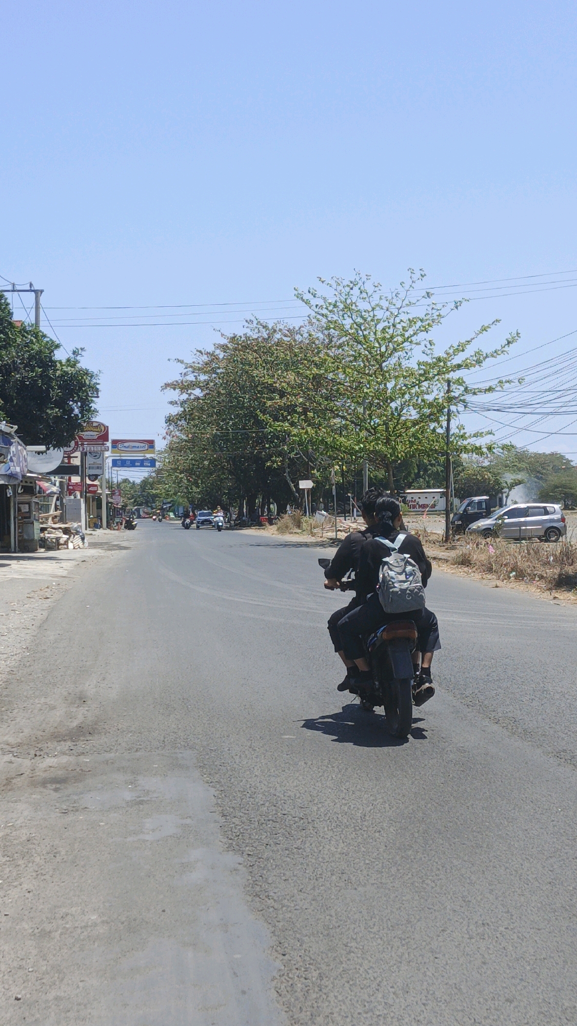 The image captures a street scene in Pangandaran, Indonesia, under a clear blue sky. Two individuals are riding a motorcycle on the right side of the road, moving away from the viewer. The person in the front is wearing black clothing, and the person in the back has a light gray backpack. The road appears to be asphalt and is in moderate condition, with some patches of lighter-colored material.
On the left side of the road, there are several small shops and buildings with awnings and signage, including what appears to be an "Indomaret" convenience store. Trees line the right side of the road, and further in the distance, more trees and some buildings are visible. Several other vehicles, including cars and motorcycles, are present on the road in the mid-ground and background. The overall environment suggests a warm, sunny day in a town or city.