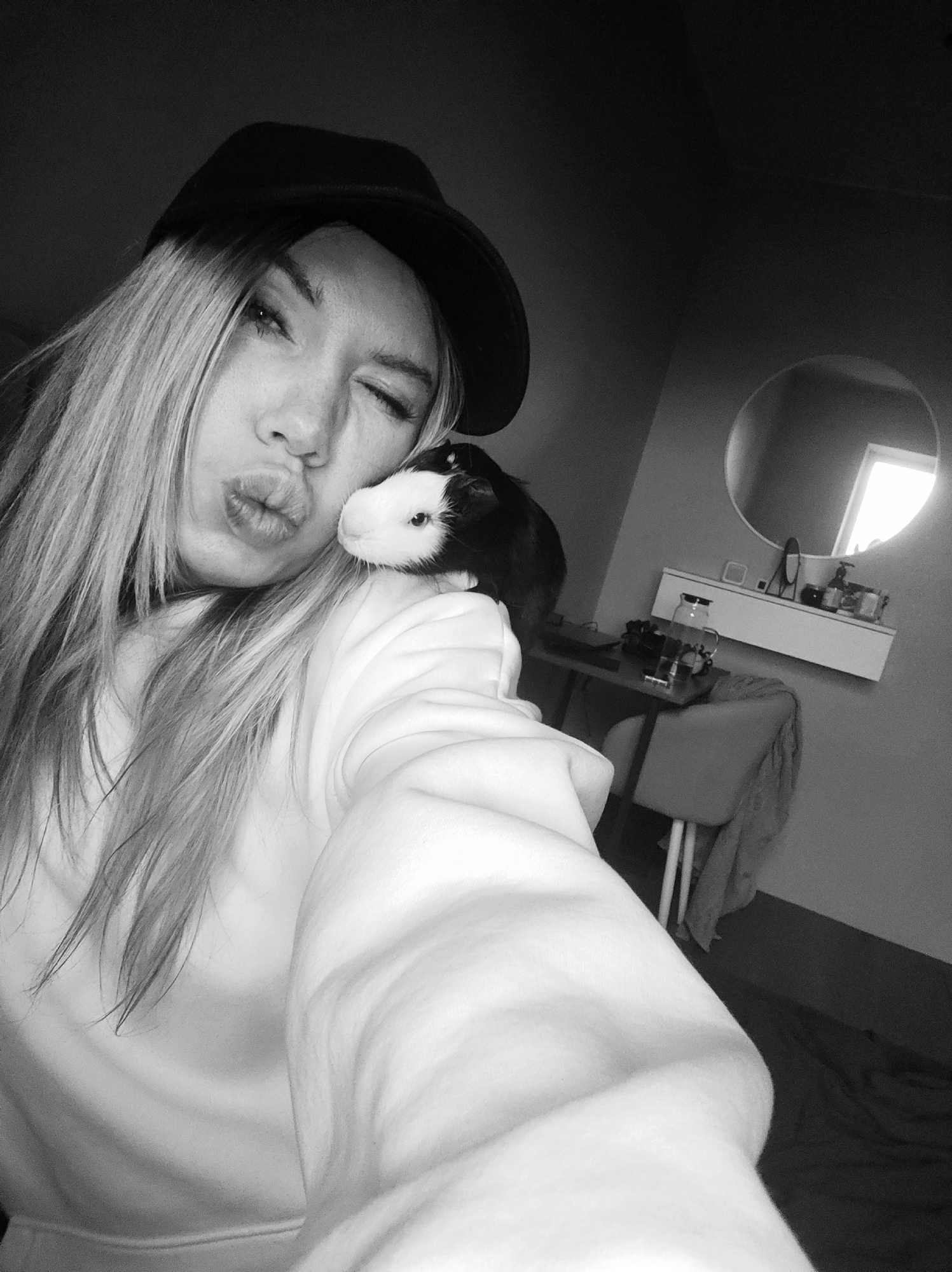 A black and white selfie captures a young woman posing playfully with a guinea pig perched on her shoulder in an indoor setting, likely a private room in Votkinsk, Russia. The woman, who has long, light-colored hair and wears a dark cap and a light-colored hoodie, is making a "kissing" face with puckered lips and has one eye winked shut. The small guinea pig, with distinctive black and white fur, is calmly positioned on her left shoulder, looking directly towards the camera.
The background reveals a room with light grey walls. A circular mirror hangs prominently, reflecting a window that suggests natural daylight entering the space. Below the mirror, a white wall-mounted shelf holds various personal items, including bottles and containers. A desk or table is positioned beneath the shelf, accompanied by a chair over which a dark cloth is draped. A clear glass pitcher or kettle and other indistinct objects are visible on the desk surface. The overall tone of the image is affectionate and casual. There is no visible text in the image.