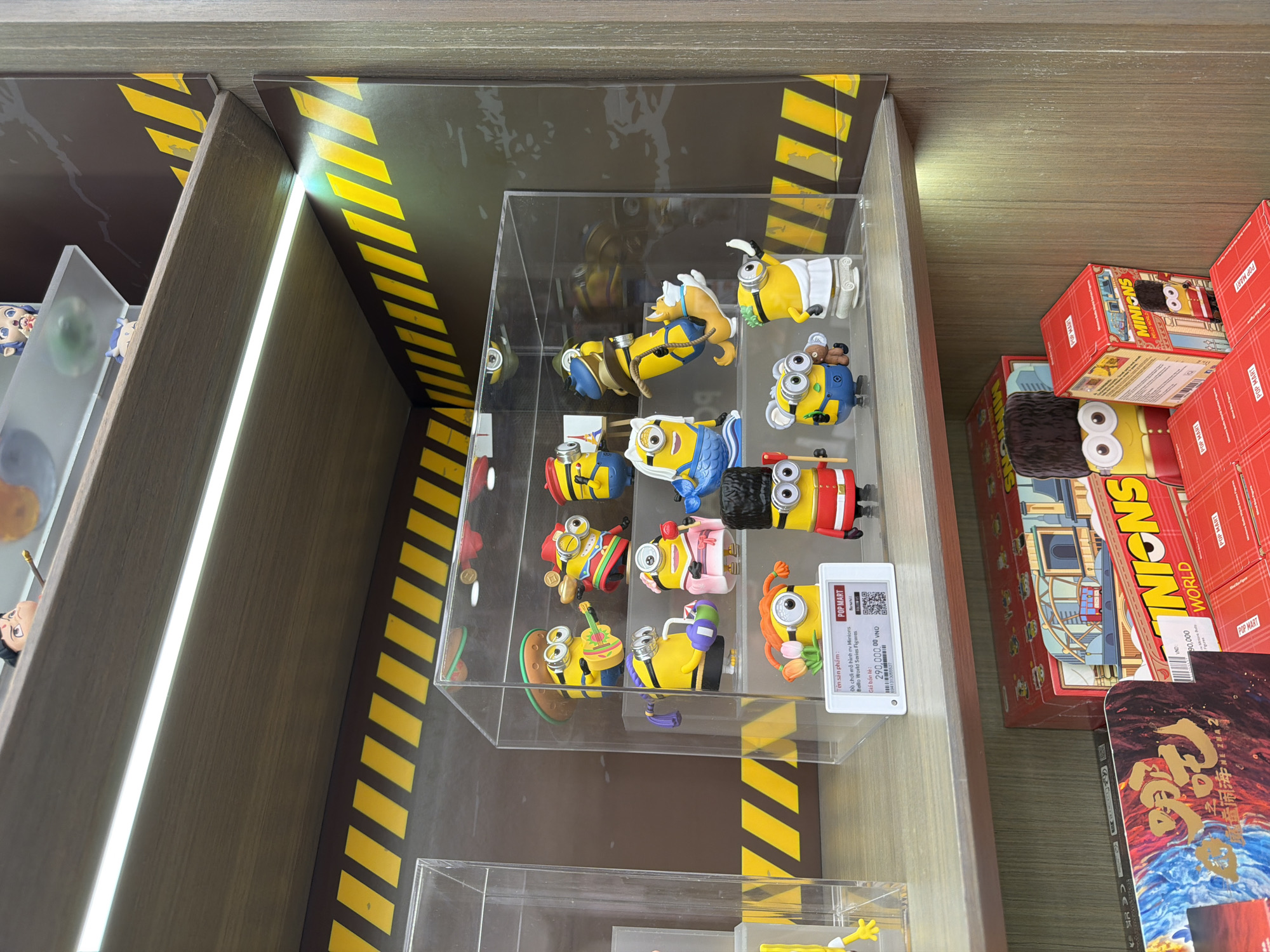The image depicts a display case filled with Minions figurines in a retail setting. The display case is made of clear acrylic and is positioned on a shelf unit with a wood-grain finish. The shelf unit has black and yellow hazard stripes on the vertical supports. Inside the display case, there are approximately twelve Minions figurines, each depicting a different character or theme.
To the right of the display case are several boxes of Minions products and other merchandise. The price tag on a Minion figurine reads "290.000,00 VND", which is Vietnamese currency, suggesting the image was taken in Vietnam. This specific price tag and the presence of the Vietnamese language on the product packaging suggest that the image was likely taken in Đà Nẵng, Vietnam.