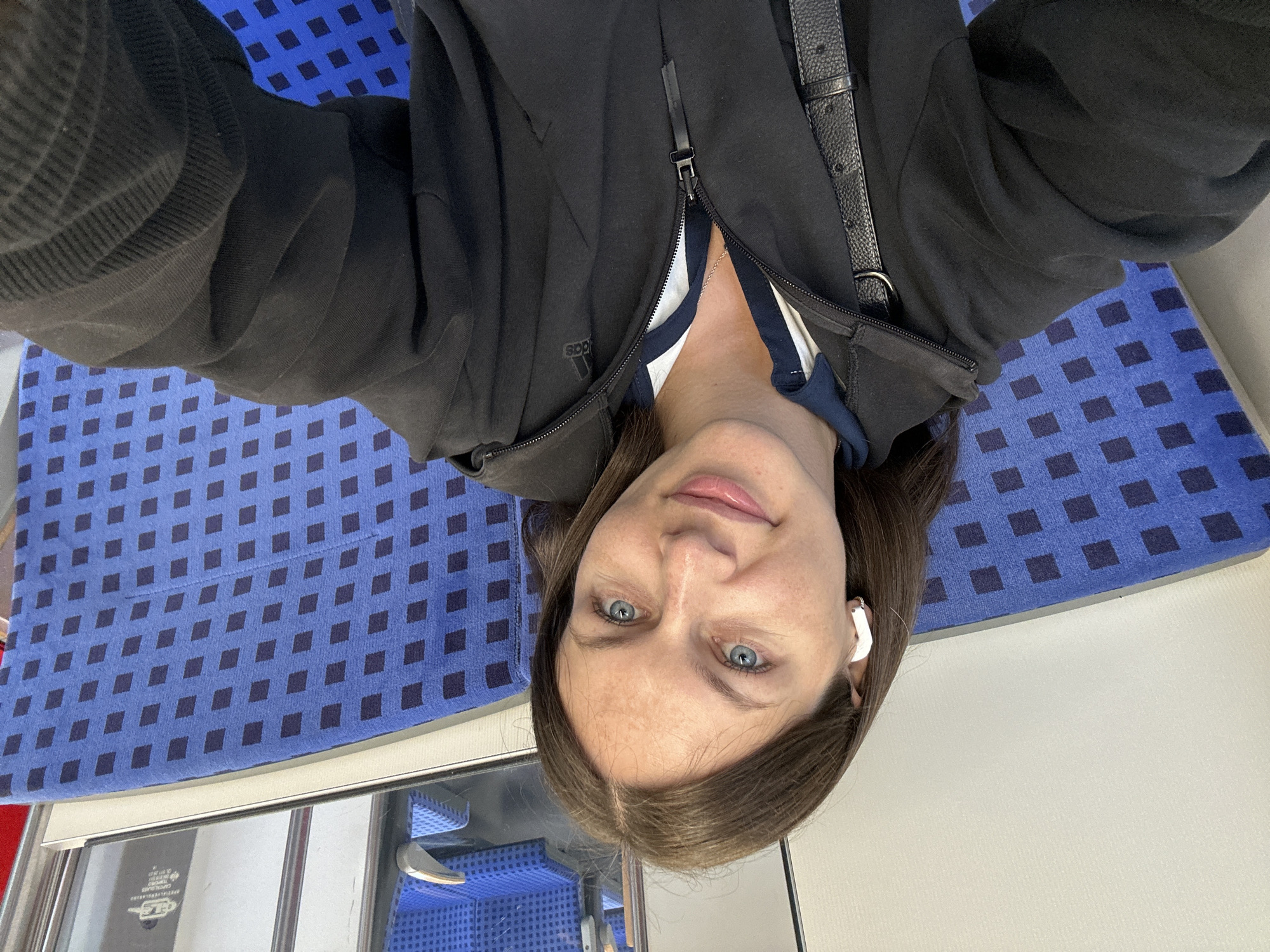 A high-angle selfie shows a woman inside what appears to be public transportation, likely a train or bus, with the image rotated 180 degrees such that her head is at the bottom. She has long, dark brown hair parted in the middle and striking blue eyes, looking directly at the camera with a subtle, calm smile. She wears white wireless earbuds in both ears.
Her attire consists of a black zippered hoodie, partially open to reveal a white and navy blue striped shirt underneath. A delicate silver chain with a small pendant is visible around her neck. The Adidas logo is present on the left side of her hoodie. A black leather strap, possibly from a shoulder bag, is visible over her right shoulder.
The background primarily features blue upholstered seats with a repeating pattern of small, dark blue diamond shapes, typical of public transport interiors. Below her head, reflective surfaces and a window are visible, showing a metallic frame and a faint reflection of the same blue patterned seating. Text reading "CAPITAL GLASS" is discernible on the window's reflection, along with other small, indistinct characters. The setting suggests she is traveling, consistent with the given location context of Buxtehude, Germany.