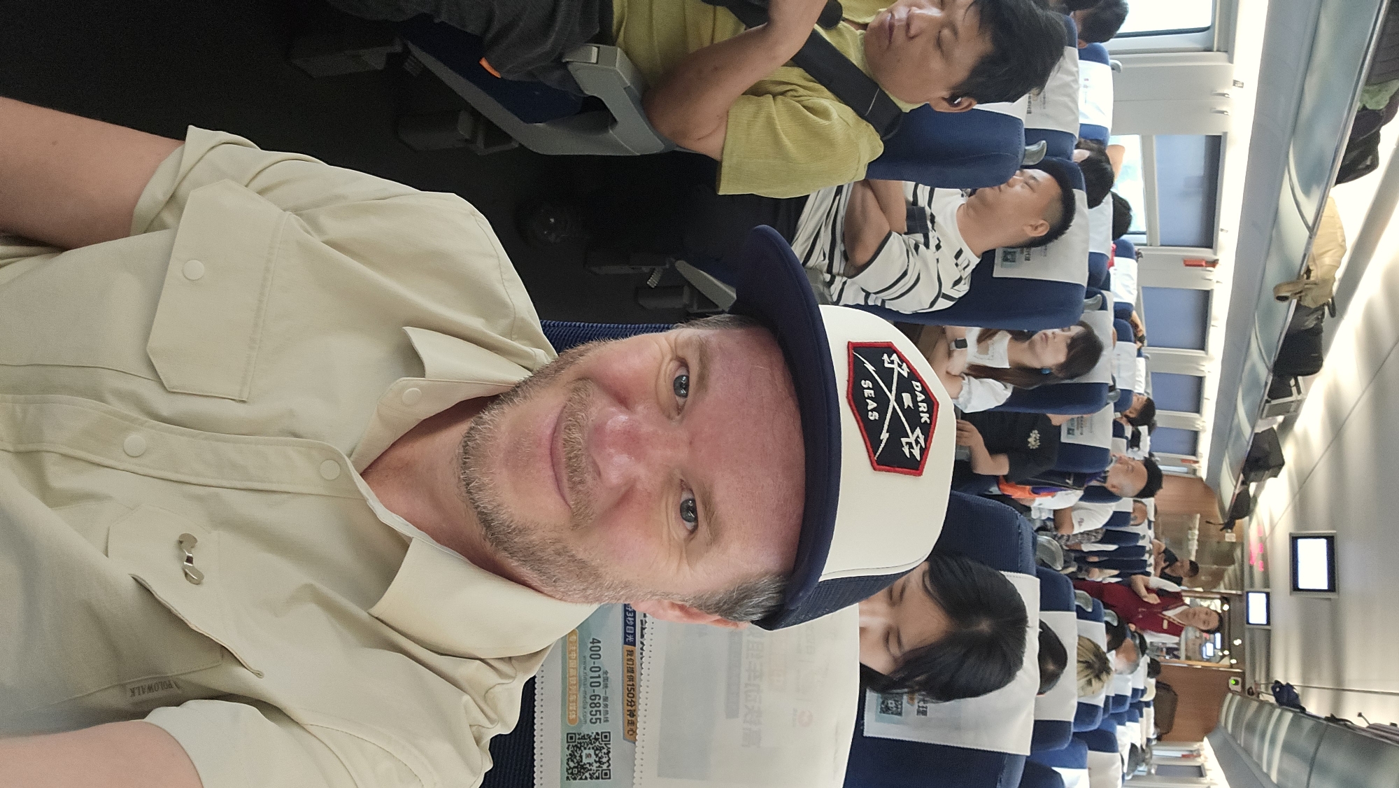 A man in a tan short-sleeved button-up shirt and a white and navy baseball cap with a black and red logo that reads "DARK SEAS" is taking a selfie on a train. The man has light skin, a light beard, and blue eyes. He is smiling at the camera. Behind him, other passengers are visible, some sleeping, some looking forward, and a flight attendant in a red uniform is standing in the aisle. The train car appears to be occupied by a diverse group of people, suggesting a public transportation setting. The seats are upholstered in blue and white. Luggage is stored in overhead compartments. The overall environment is that of a busy train journey.