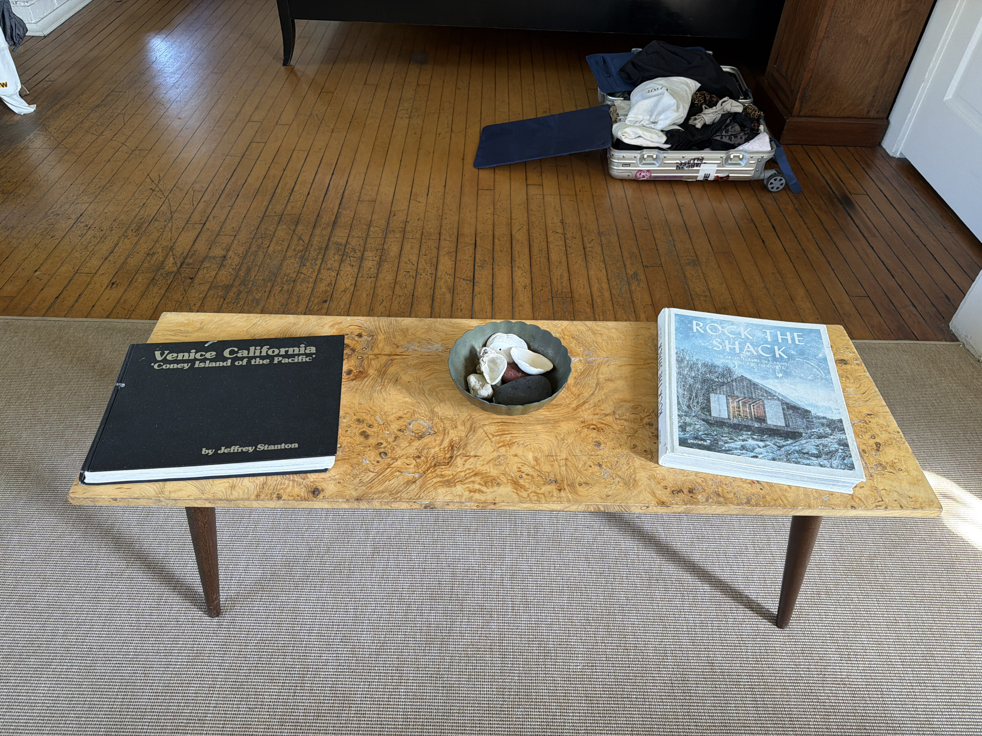 An indoor, eye-level shot captures a domestic scene featuring a coffee table, books, and an open suitcase. In the foreground, a light-colored, tightly-woven area rug covers the floor. Centered on the rug is a mid-century modern-style coffee table with a light, burl wood top and four dark, tapered legs.
On the coffee table, two large books are displayed. To the left is a black-covered book titled "Venice California 'Coney Island of the Pacific'" by Jeffrey Stanton. To the right is a light-colored book titled "ROCK THE SHACK" with "THE ARCHITECTURE C" and "gestalten" also visible. Between the books, a small, dark metal bowl holds several white seashells and dark stones.
Behind the coffee table, a worn, light brown hardwood floor stretches into the background, reflecting bright light from an unseen window, suggesting it is daytime. On the floor in the upper right, an open, silver-colored suitcase is packed with clothes, including a white t-shirt with "DIOR" visible and some leopard print fabric. A blue, rectangular mat is partially visible under the suitcase. To the far right, a white door frame and part of a dark wooden furniture piece are seen. In the upper left, a small portion of a white object with the letter "W" is visible.
The scene depicts a lived-in space, possibly in a living room or study, within Los Angeles, United States of America. The open suitcase suggests travel or recent unpacking.