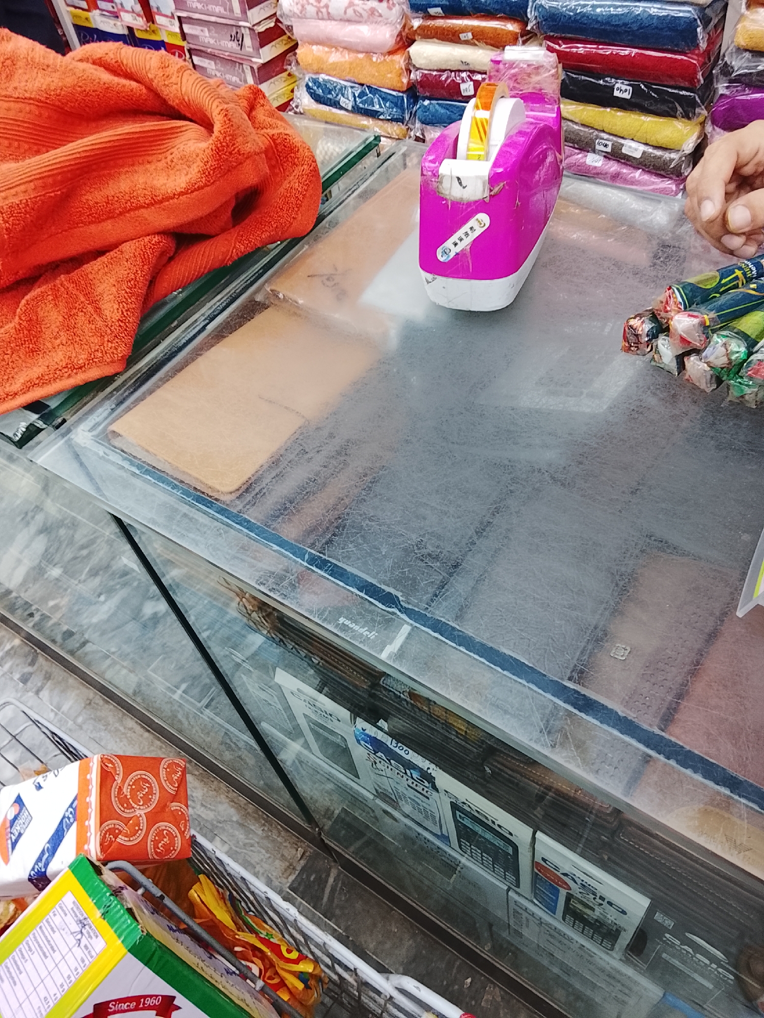 The image depicts a commercial environment, likely a retail establishment in Gulgasht Colony, Pakistan. On a glass countertop, an orange towel is folded to the left of the frame. A pink tape dispenser sits beside a collection of wrapped cylindrical objects. Behind these items, a hand is visible. Behind and above the counter items are various items packaged in cellophane and stacked. Visible under the glass countertop are boxes of calculators. The light suggests an indoor setting.