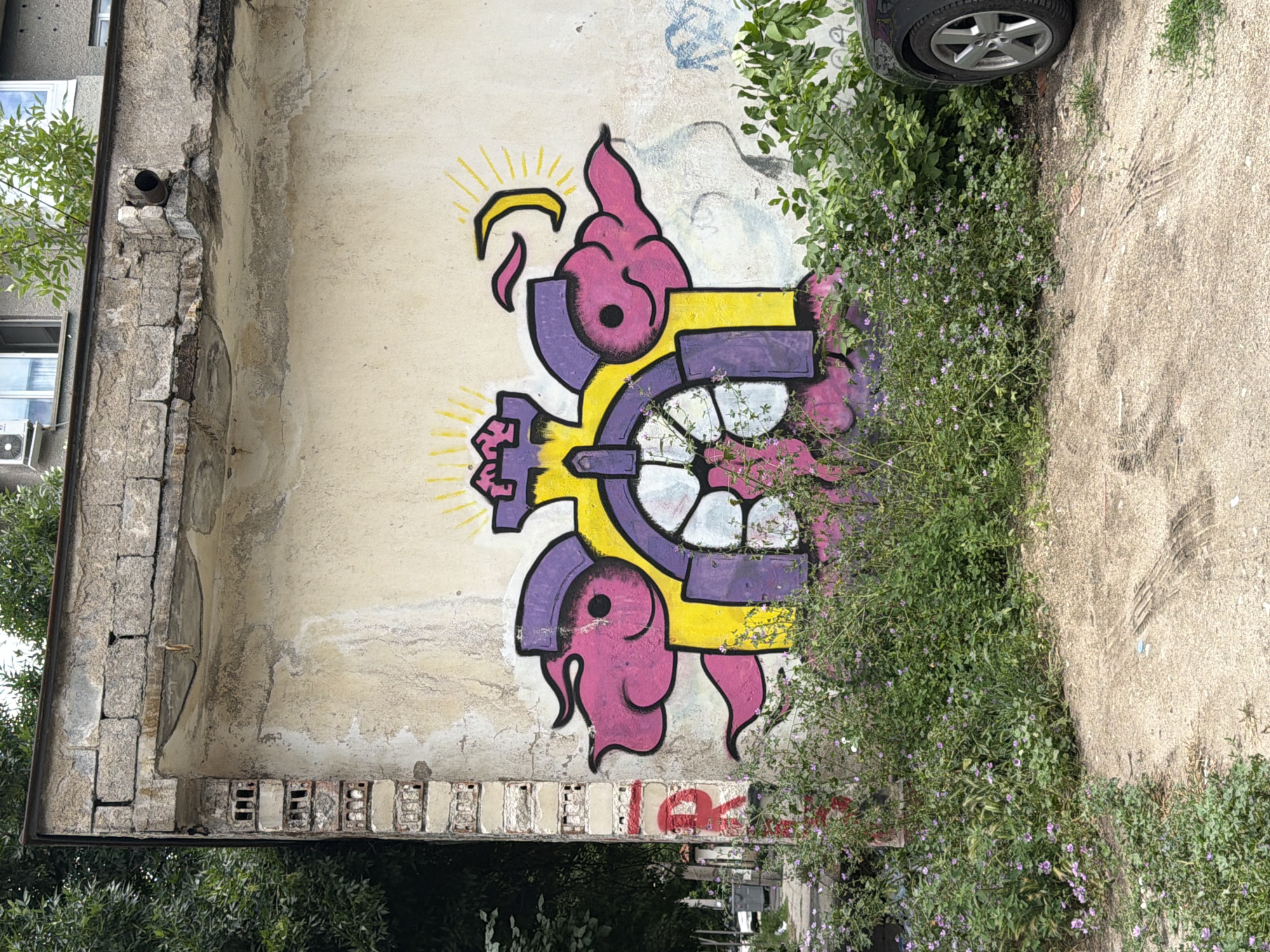 The image shows a section of an exterior wall of a building in Pazardzhik, Bulgaria, featuring graffiti. The wall is weathered and stained, with signs of disrepair, including crumbling brickwork along the top edge and a rusting pipe protruding from it. The graffiti itself is colorful and cartoonish, depicting a yellow and purple abstract design that resembles a stylized sun or clock with pink cloud-like elements on either side and a crescent moon shape above. The ground in front of the wall is a mixture of dirt and concrete, with tire tracks visible in the dirt. Overgrown green vegetation and small purple flowers are growing in front of the graffiti and along the base of the wall. A portion of a black car is visible on the right side of the image, parked on the concrete. In the background, trees and a portion of another building with windows and an air conditioning unit can be seen.