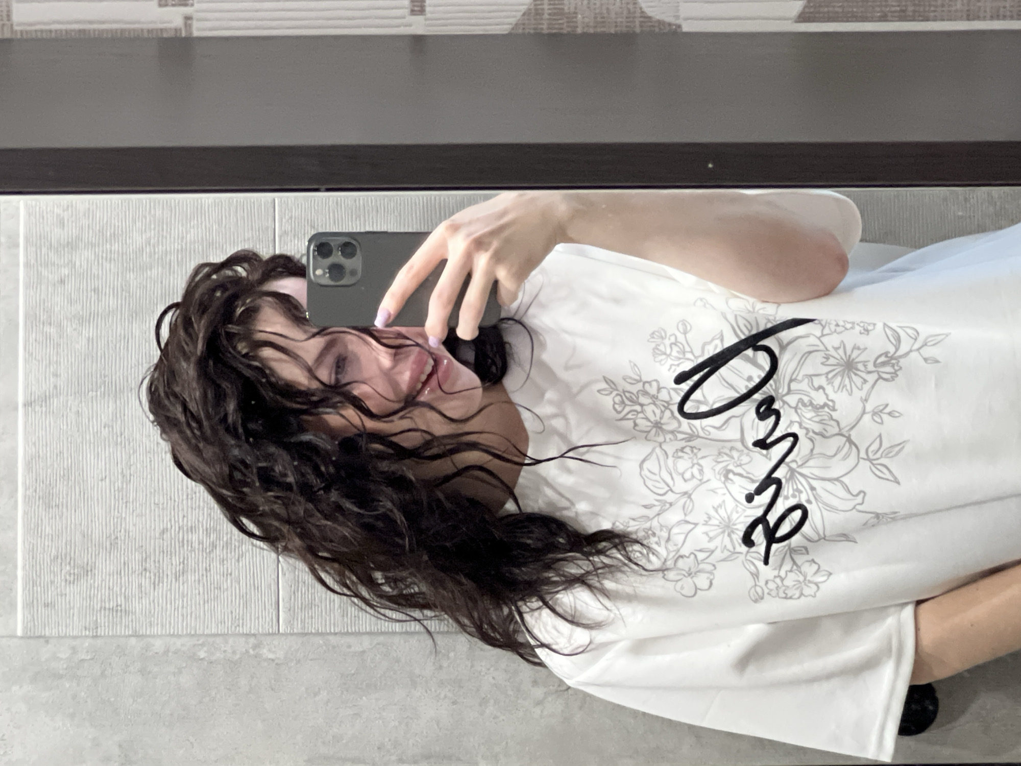 A woman with long, dark, wet hair is taking a selfie. She is holding a smartphone with multiple cameras, and her fingers are visible. She is wearing a white t-shirt with a floral design and the word "Love" in cursive script. The background consists of a tiled wall and a dark countertop. The location appears to be a bathroom interior, with tile flooring. There is no information available to confirm this photo was taken in Krasnodar, Russia.