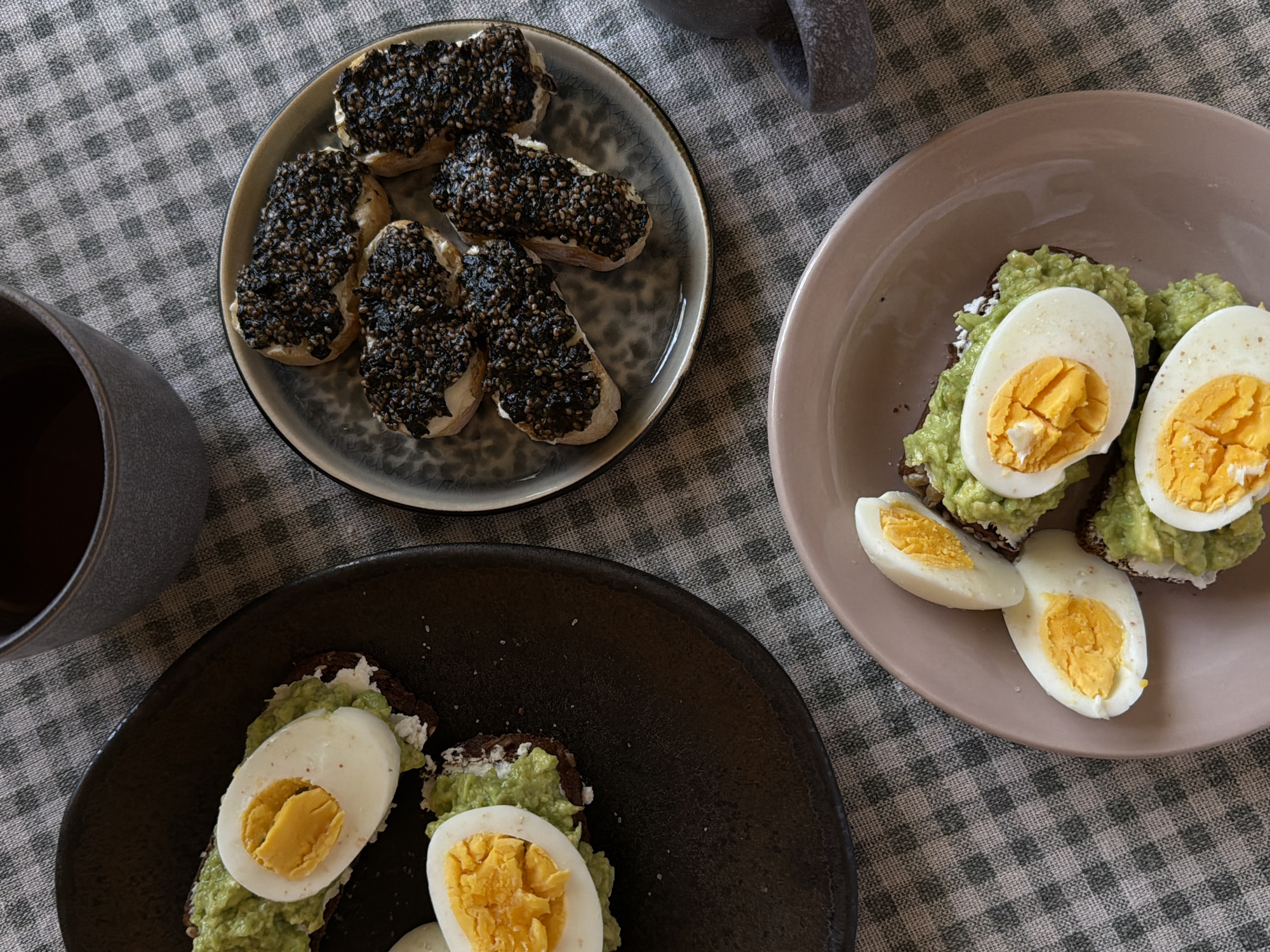 The scene presents a meal of open-faced sandwiches on a checkered tablecloth. In the upper left, a plate contains small bread slices covered in a dark, granular spread, possibly black sesame seeds. A dark-colored mug sits nearby.
To the right, two plates hold avocado toast topped with sliced hard-boiled eggs. One plate is a light brown, and the other is dark brown. The toast appears to be on dark bread. A mortar and pestle are partially visible in the upper right corner.
The lighting is soft and natural, suggesting daytime. The context indicates a casual meal setting, perhaps breakfast or brunch. There is no evidence to indicate this scene is taking place in Khimki, Russia.