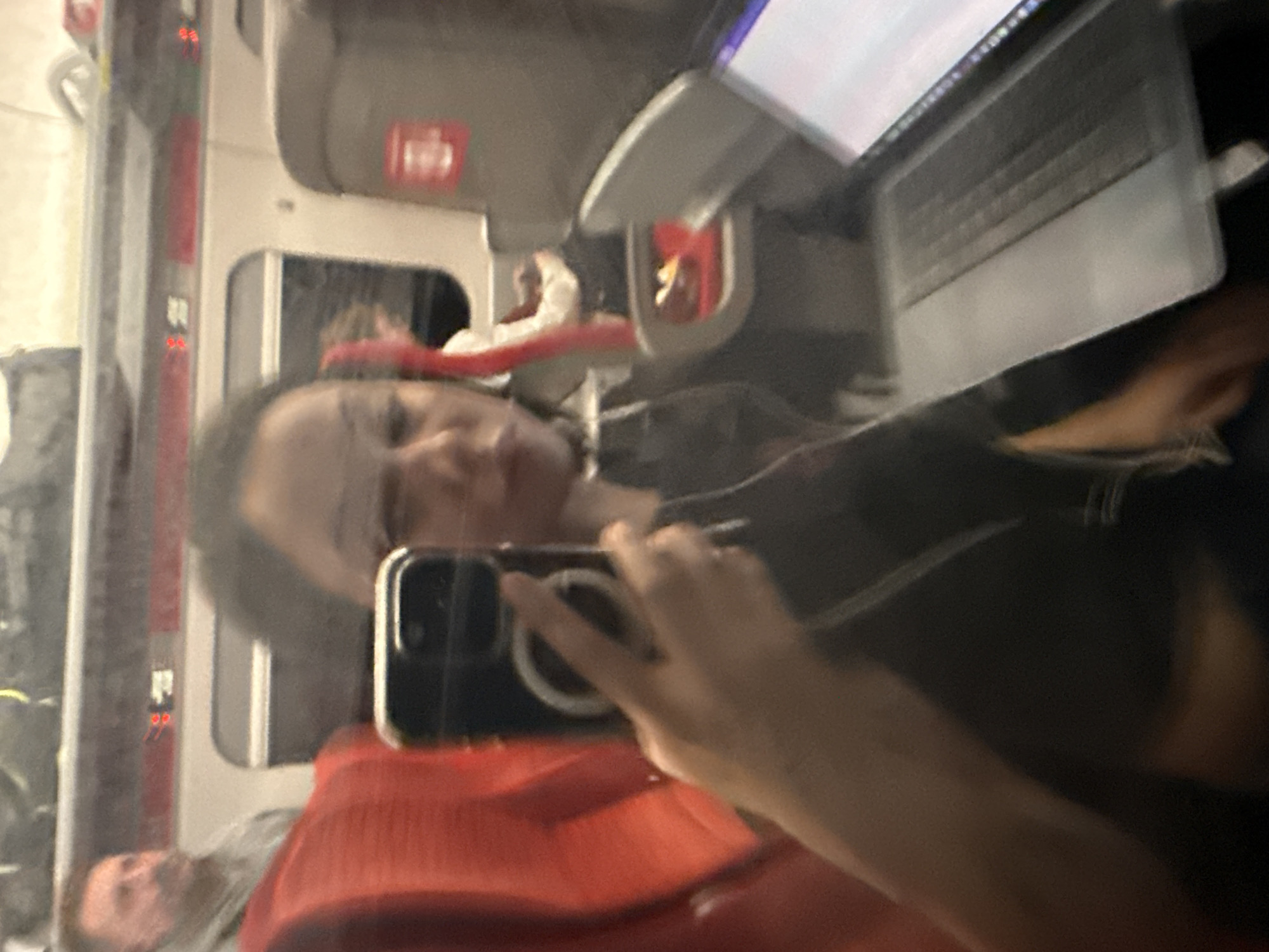 A woman is taking a selfie in what appears to be the interior of a train. She is holding a smartphone in her right hand, capturing her reflection in a window. She is wearing a red garment and appears to be positioned near a laptop. Other passengers are visible in the background.
The train's interior shows features such as overhead storage and seating arrangements. The city of South Kesteven is not visible in this photograph.