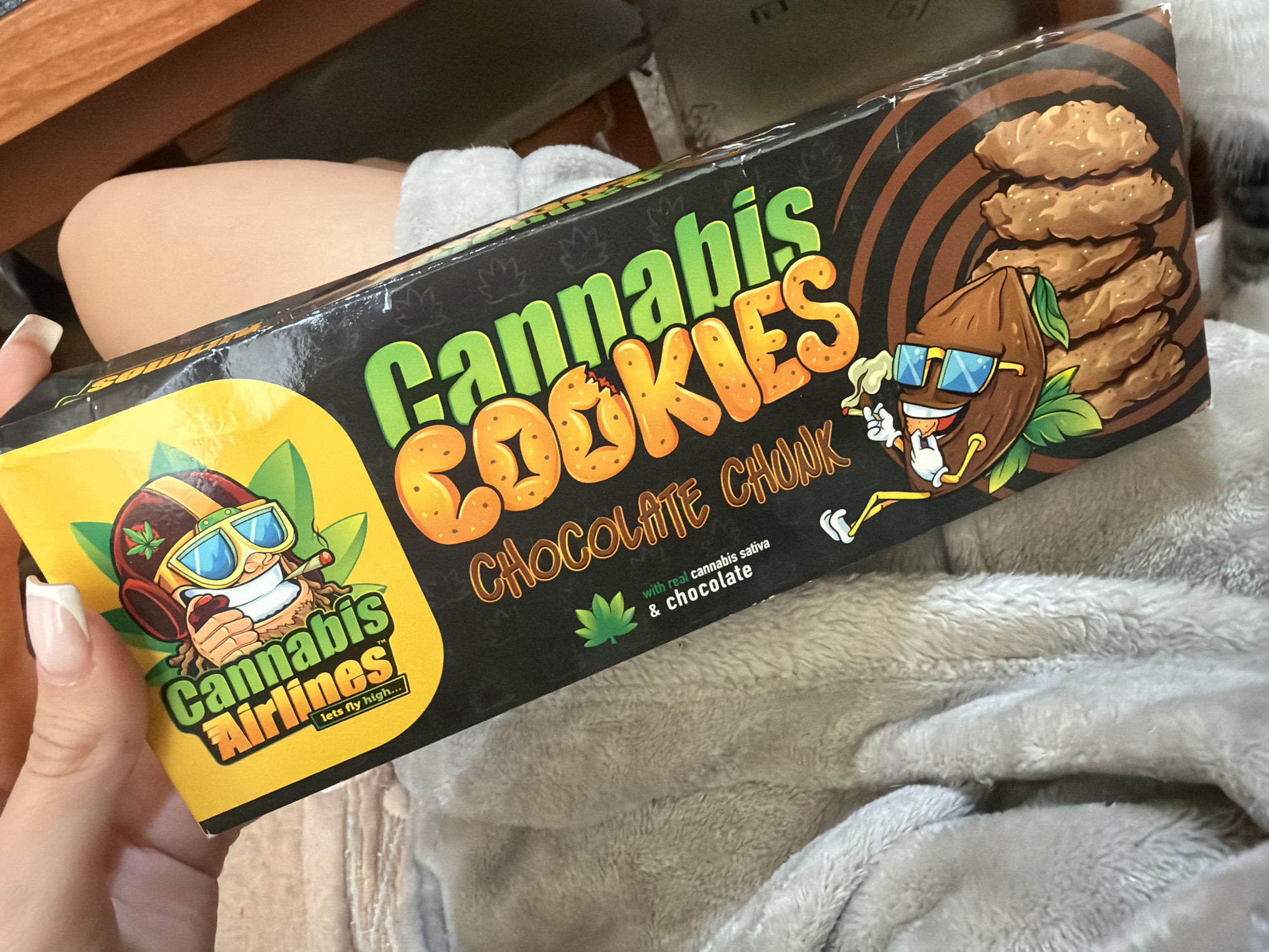 The image captures a person's hand holding a rectangular box of "Cannabis Cookies" in an indoor setting in Burgas, Bulgaria. The product box is prominently displayed, featuring a black background with vibrant green and orange lettering for "Cannabis COOKIES" and brown text for "CHOCOLATE CHONK." Cartoon illustrations are central to the packaging design: one depicts a smiling brown character, resembling a cacao bean or seed, wearing blue sunglasses and smoking a joint, positioned next to a stack of chocolate chip cookies. Another character, similar in style but wearing a pilot's helmet and goggles, also smoking a joint, appears on a yellow panel with the "Cannabis Airlines" logo and the tagline "lets fly high...". The box explicitly states "with real cannabis sativa & chocolate," accompanied by a green cannabis leaf symbol, which is also subtly patterned across the black areas of the box.
The hand holding the box has manicured nails with a white French tip. Below the box, a soft, light grey, textured blanket or throw covers a person's lap or legs, with a glimpse of bare skin (likely a knee or thigh) visible beneath it. In the upper background, part of a brown, wooden surface, possibly a table or desk, can be seen. The image is a close-up, focusing on the product packaging and the hand holding it.