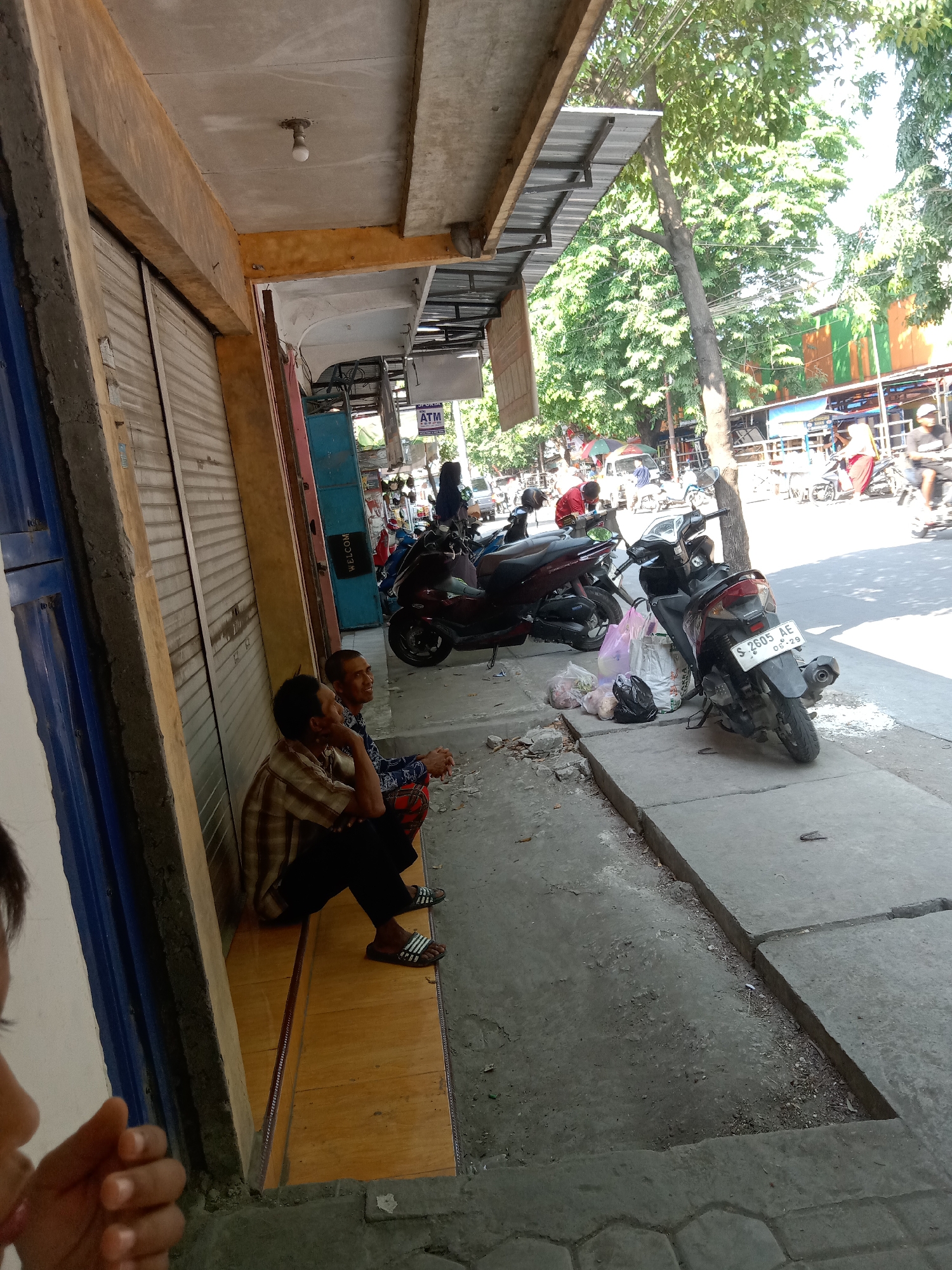 Two men are sitting on a yellow wooden platform adjacent to a closed storefront in Sugihwaras, Indonesia. The storefront has a corrugated metal roll-down gate. The man on the left is wearing a checkered shirt and dark pants, and is looking to his right. The man on the right is wearing a blue and white patterned shirt and plaid pants, and is looking to his left. They are sitting on a sidewalk in front of shops. Several motorcycles are parked on the sidewalk and street. In the background, there are trees and other shops. The image appears to have been taken during daylight.