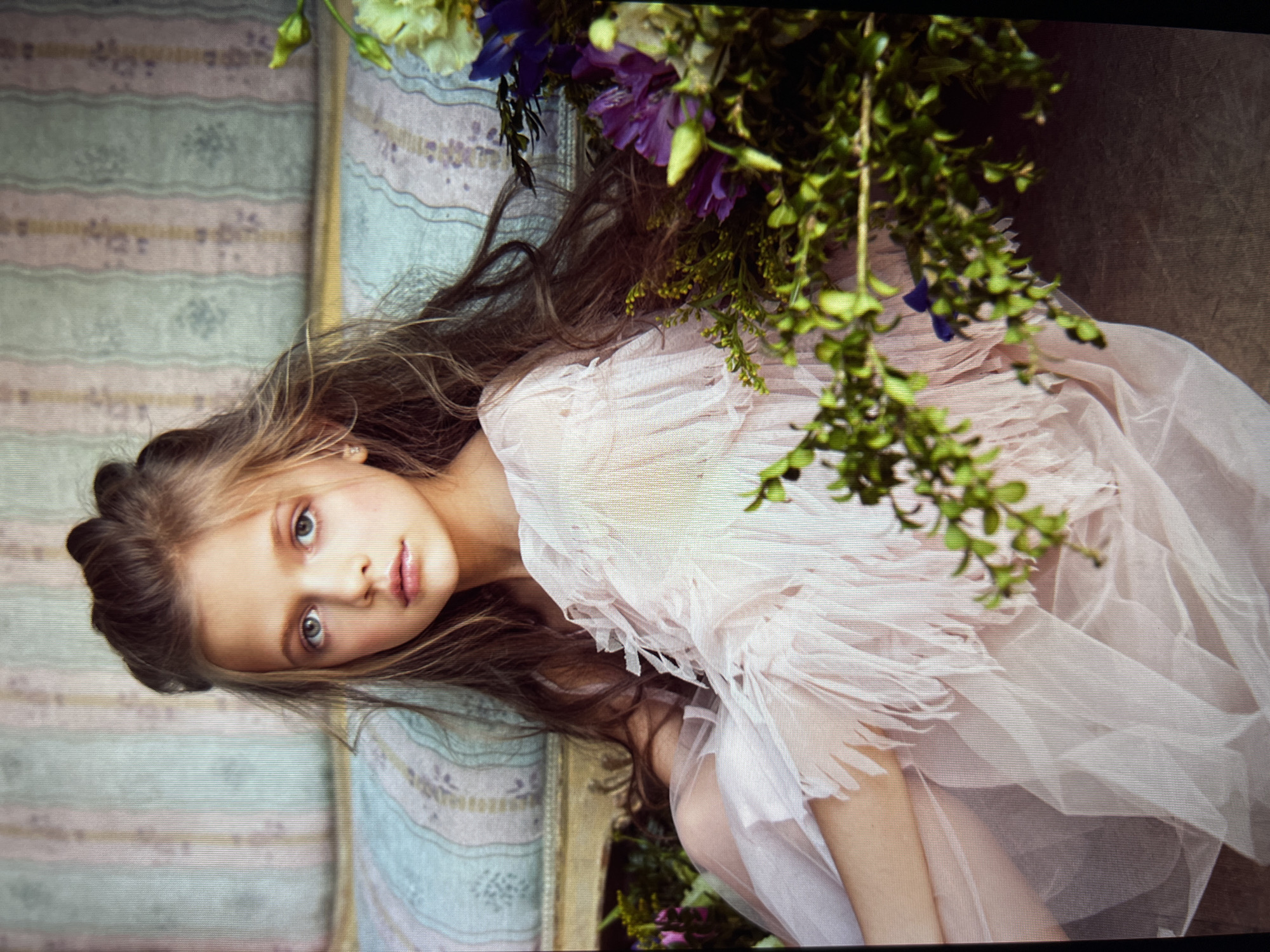 The image shows a young girl with long, brown hair lying down, her head resting on a light-colored, patterned surface. She is wearing a delicate, pale pink dress that appears to be made of layered, translucent fabric. Her eyes are wide open and looking directly at the camera with a calm expression. Her gaze is intense and captivating.
In the upper right portion of the image, there is a lush arrangement of greenery and flowers, including purple and light green blossoms, which partially drapes over the girl and her surroundings. The background to the left of the girl consists of a striped fabric in shades of pale blue, pink, and white, with subtle floral patterns. To the right of the girl, the background appears to be a darker, textured surface, possibly wood or fabric.
The lighting in the image is soft and diffused, creating a gentle, almost ethereal mood. It is difficult to determine the time of day or weather conditions from the image, but the overall aesthetic suggests a staged portrait or editorial photograph. There is no visible text in the image. The girl's pose and the presence of the floral arrangement suggest a theme of nature, beauty, or perhaps a fairy tale.