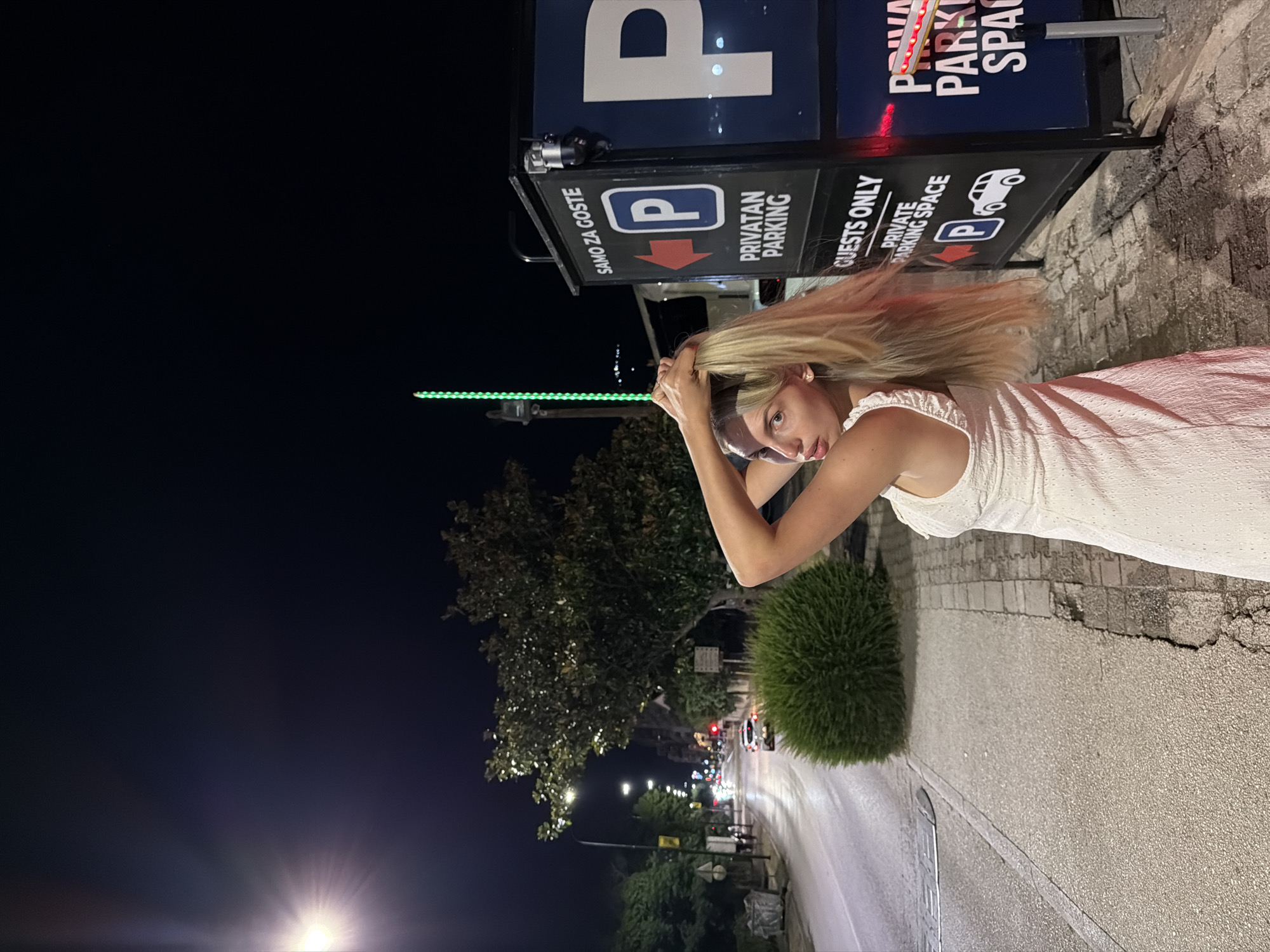 A young woman with blonde hair, wearing a white dress, stands on a sidewalk at night. She is touching her hair and looking at the camera. Behind her, there is a street with cars, trees, and buildings. To her right, there is a blue sign that says "SAMO ZA GOSTE PRIVATAN PARKING GUESTS ONLY PRIVATE PARKING SPACE". The scene appears to be in the city of Budva, Montenegro, as indicated by the text on the parking sign which is in Croatian.
